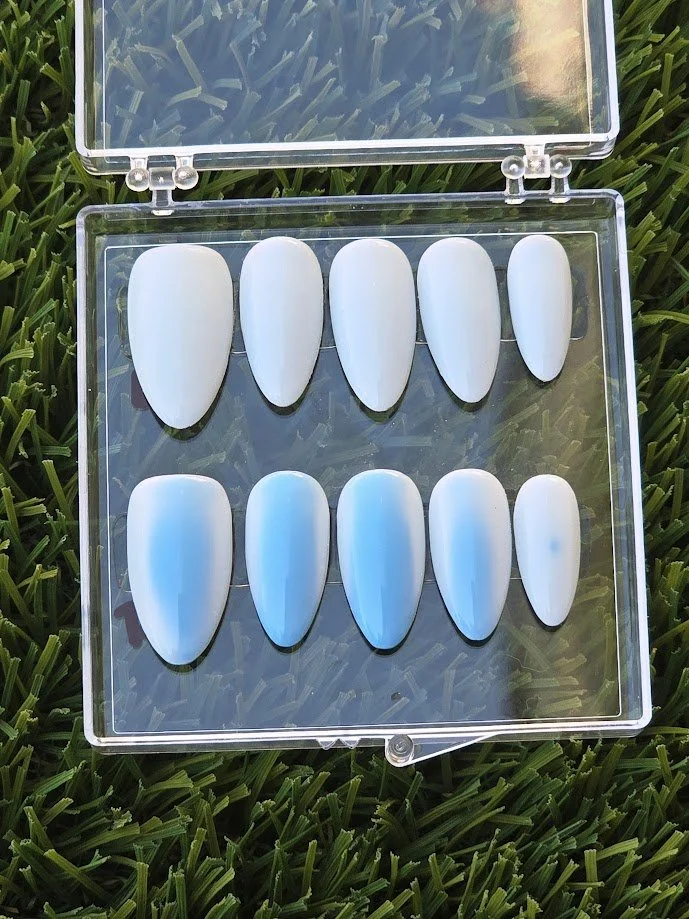 A clear plastic case containing ten artificial nails on a grassy background, with five nails in two rows, all painted in an ombre white to blue gradient.