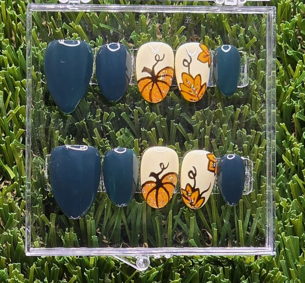 Set of ten decorative artificial nails with autumn and pumpkin-themed designs, arranged in two rows inside a clear display box, on a background of green grass.