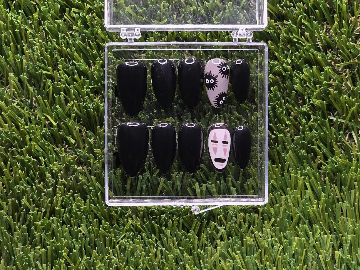 A set of decorative black and printed press-on nails arranged in a clear plastic case, placed on green grass.
