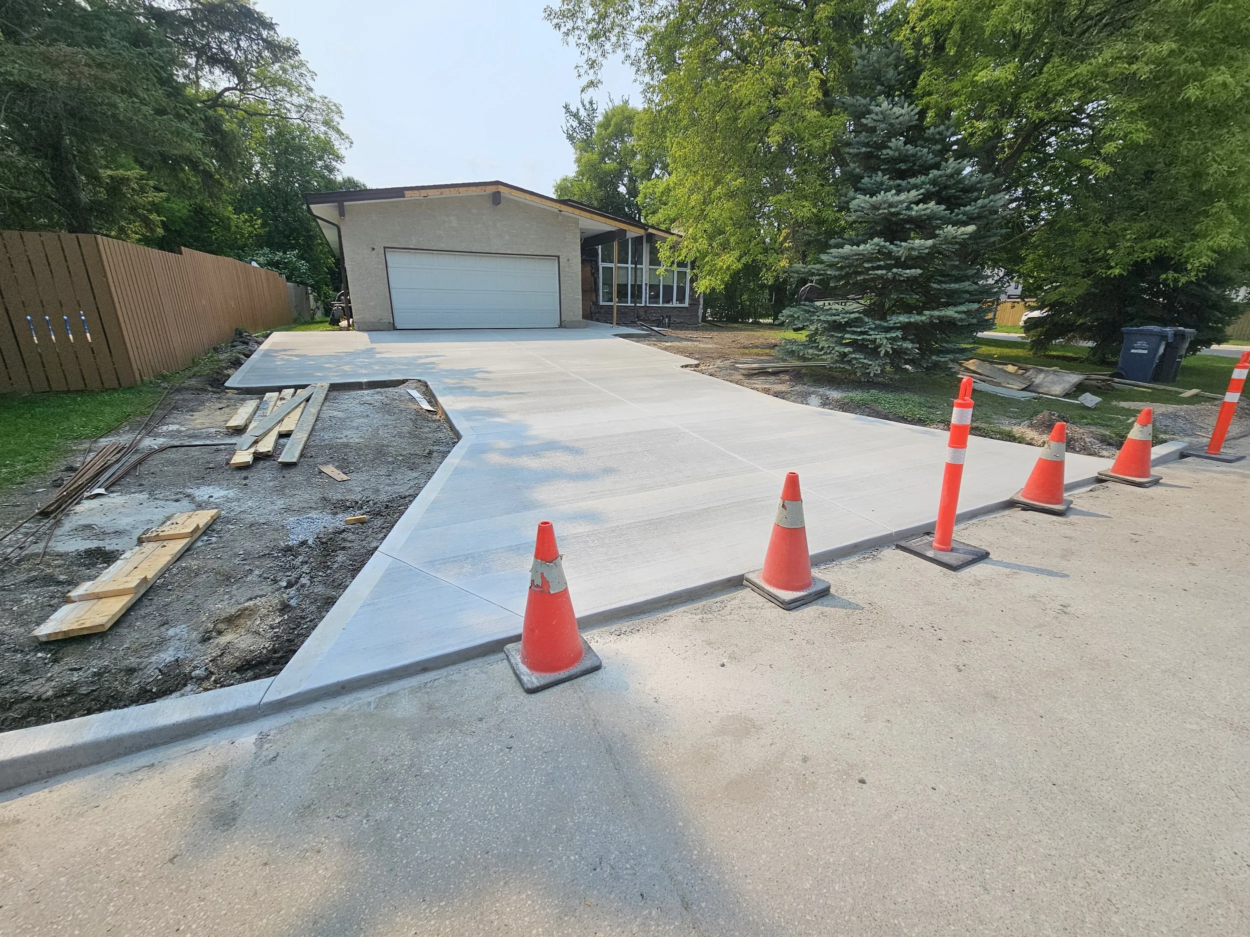 Newly poured concrete driveway with orange traffic cones along the edge, in front of a house with a garage, surrounded by trees and a wooden fence.