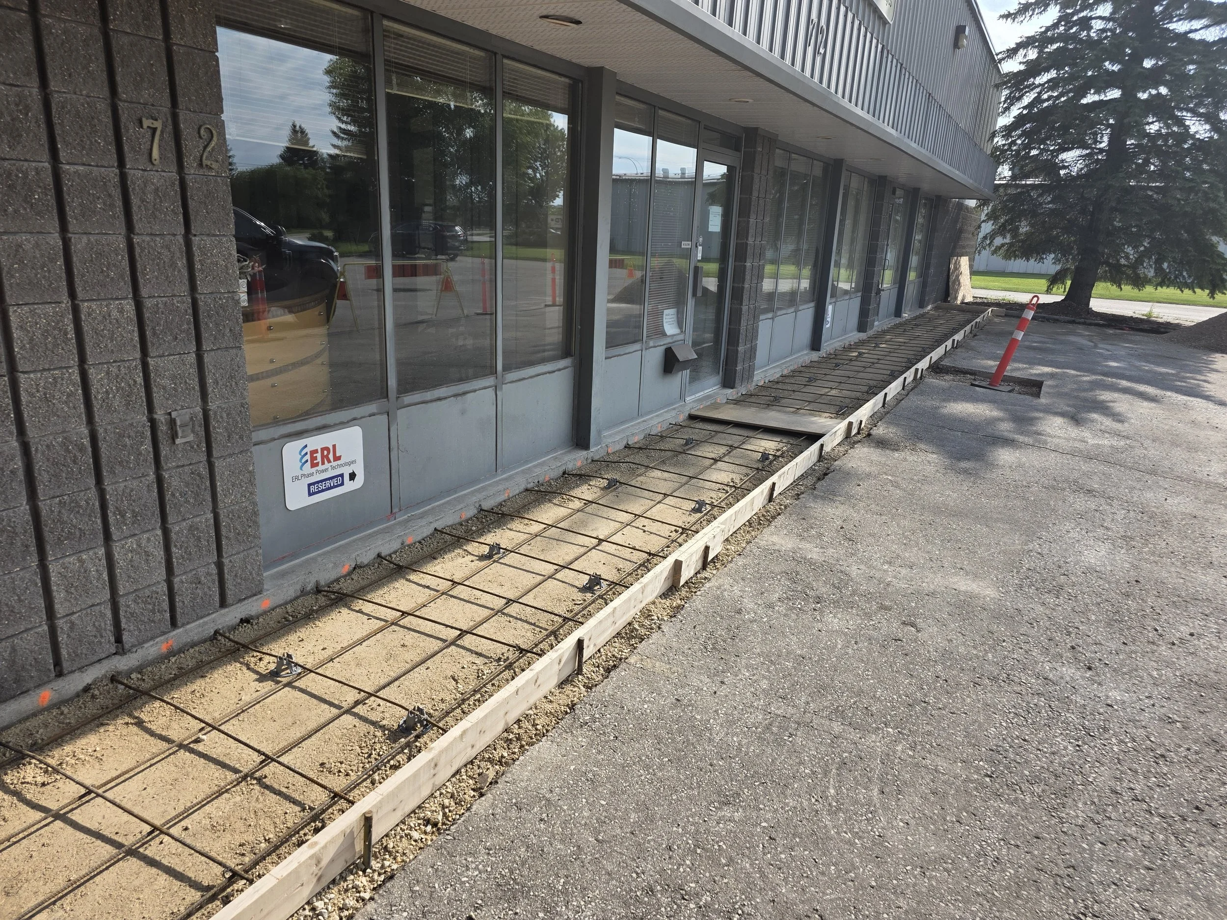 Construction work outside a commercial building with large windows, a reserved parking sign, and a sidewalk under construction with exposed rebar and wooden forms.