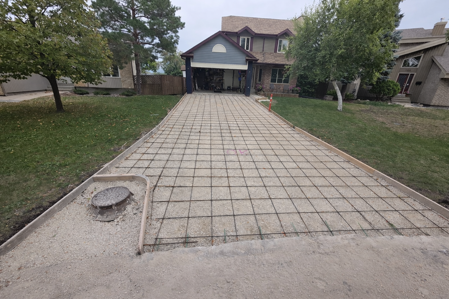 Driveway under construction with concrete paving, metal rebar grid, and wooden borders leading to a house with an open garage, trees on both sides, and neighboring houses in the background.
