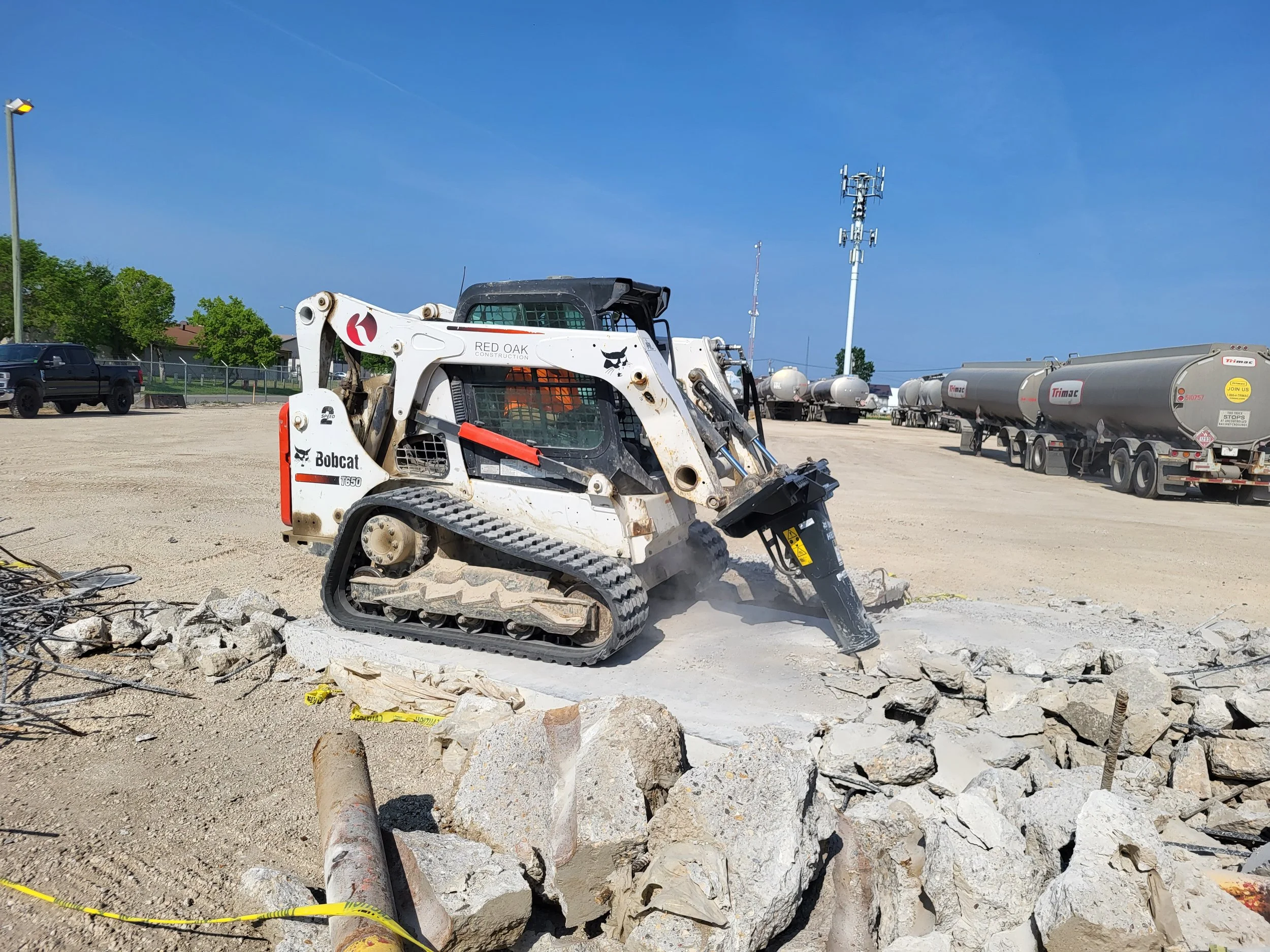 A small Bobcat excavator breaking apart concrete at a construction site with large rocks and debris in the foreground and tanker trucks in the background under a clear blue sky.