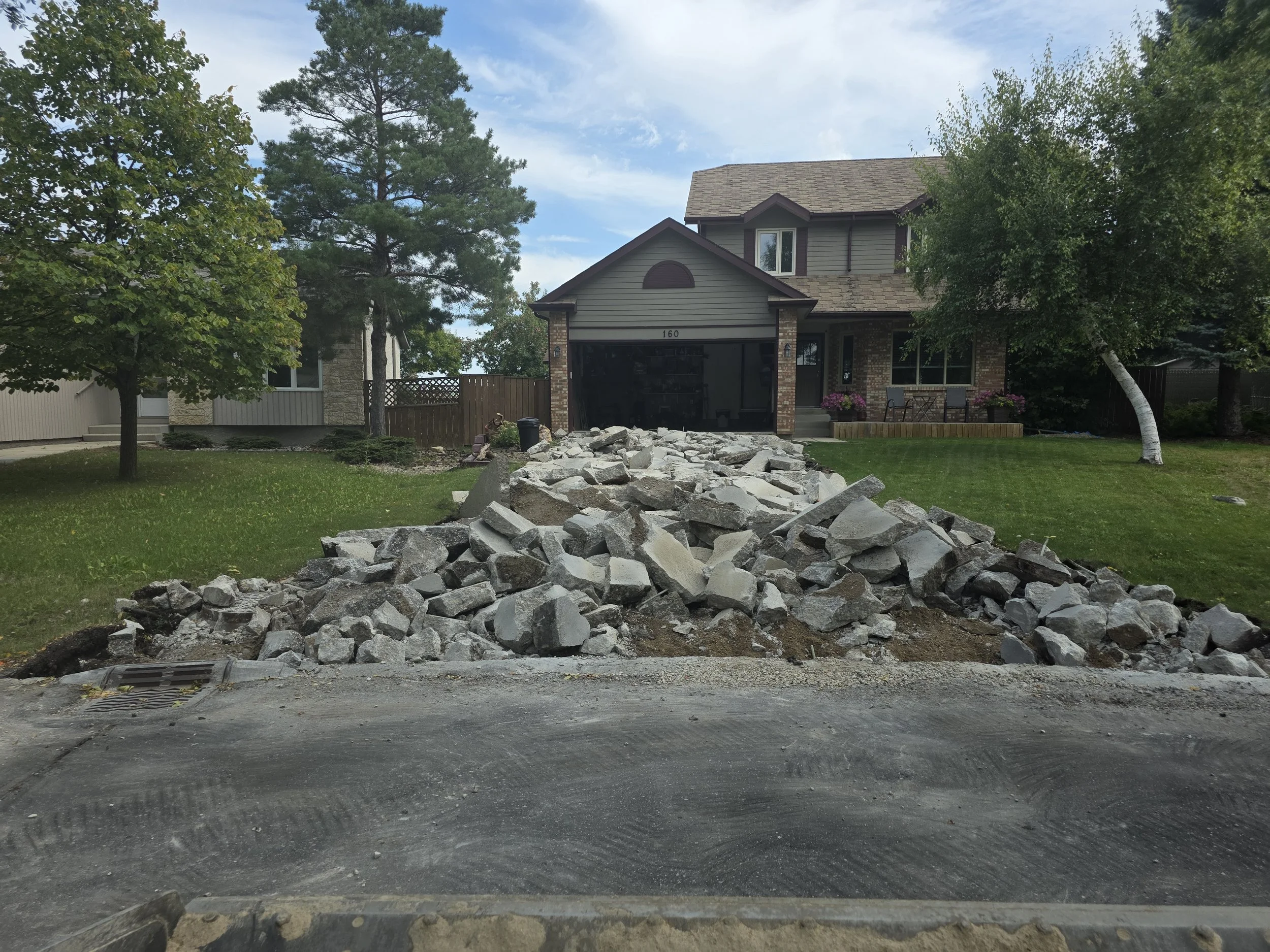 Residential street with construction debris, large rocks, and dirt in the foreground. House with brick and gray siding, trees, and a manicured lawn in the background.