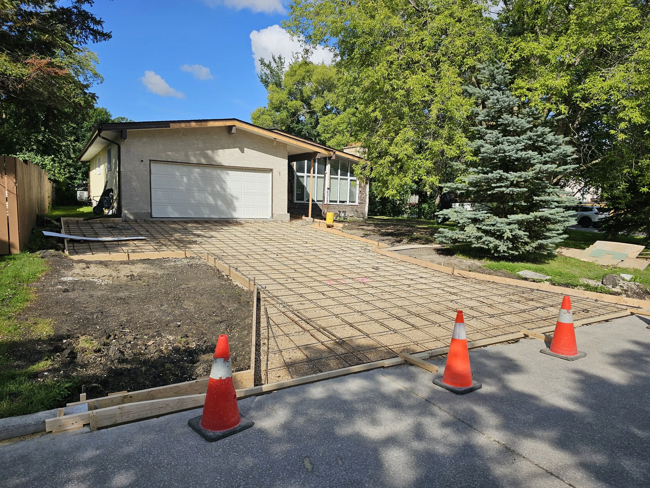 Driveway under construction with wooden framing, metal rebar, and orange traffic cones in front of a house with a garage and trees.