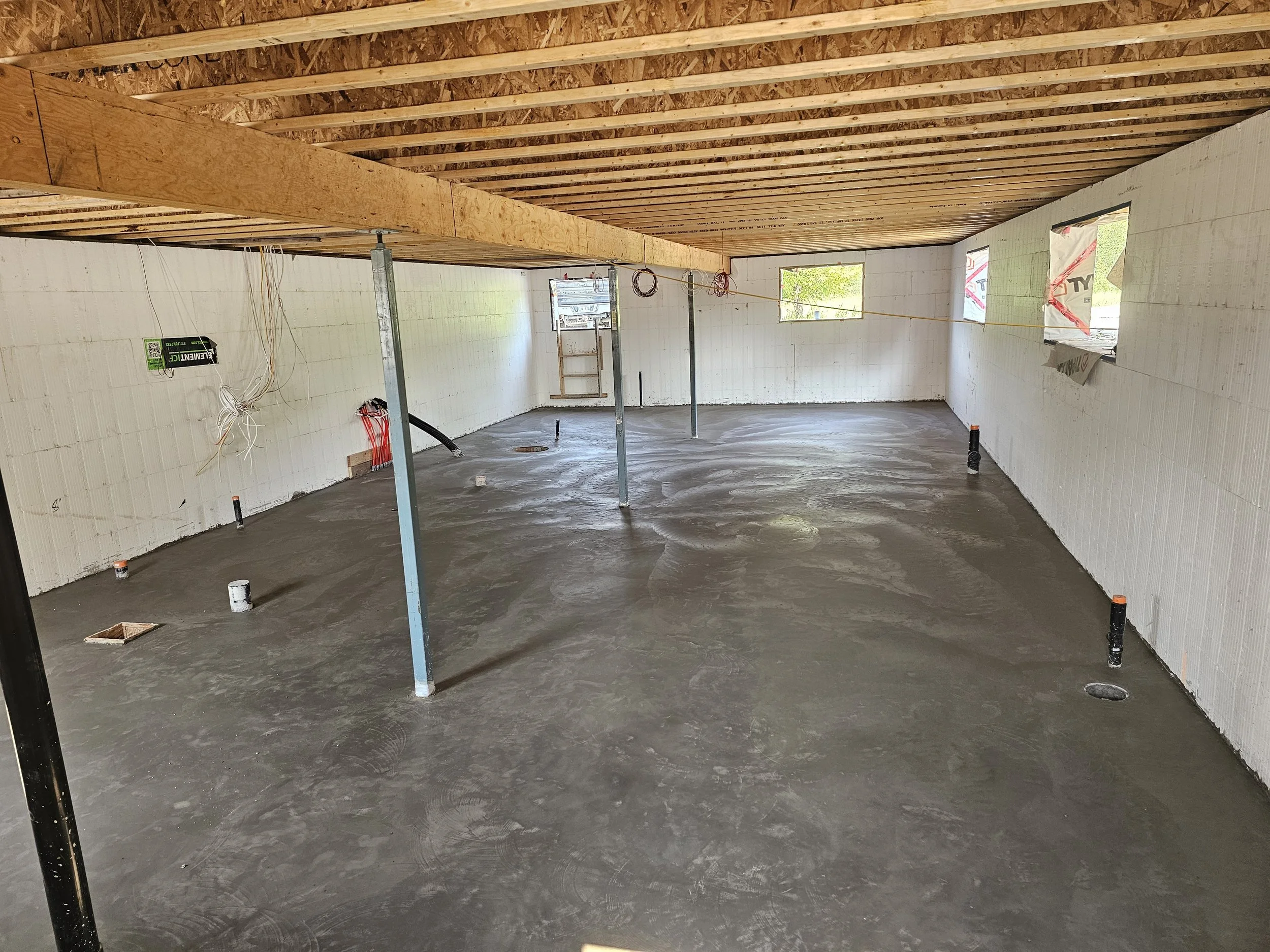 Interior of a house under construction with a concrete floor, white walls, and an exposed wooden ceiling. Support poles and construction materials are visible.