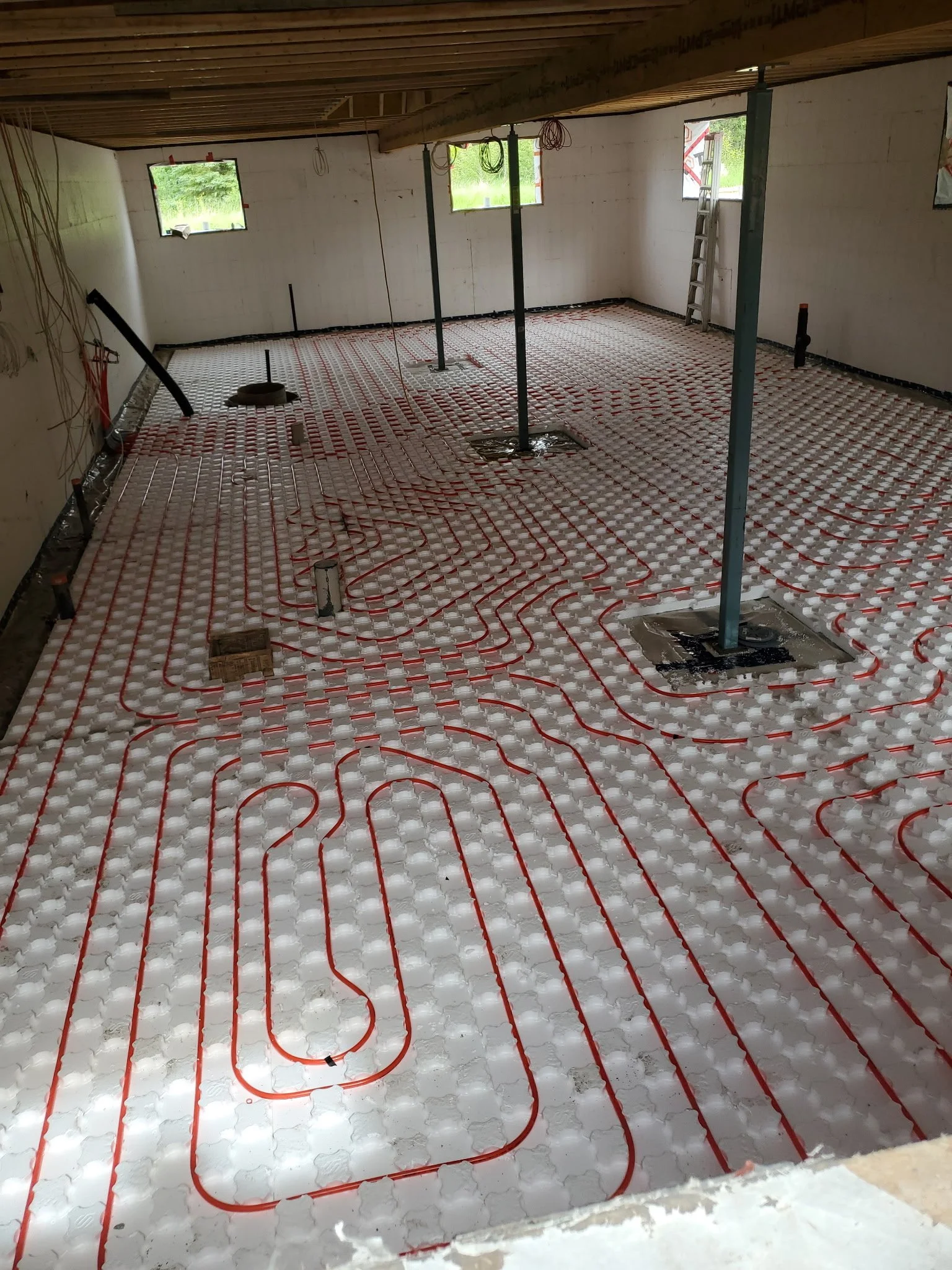 Interior view of a building under construction with red radiant floor heating pipes installed on a foam board insulation, supported by vertical steel posts, with small windows showing a green outdoor area.