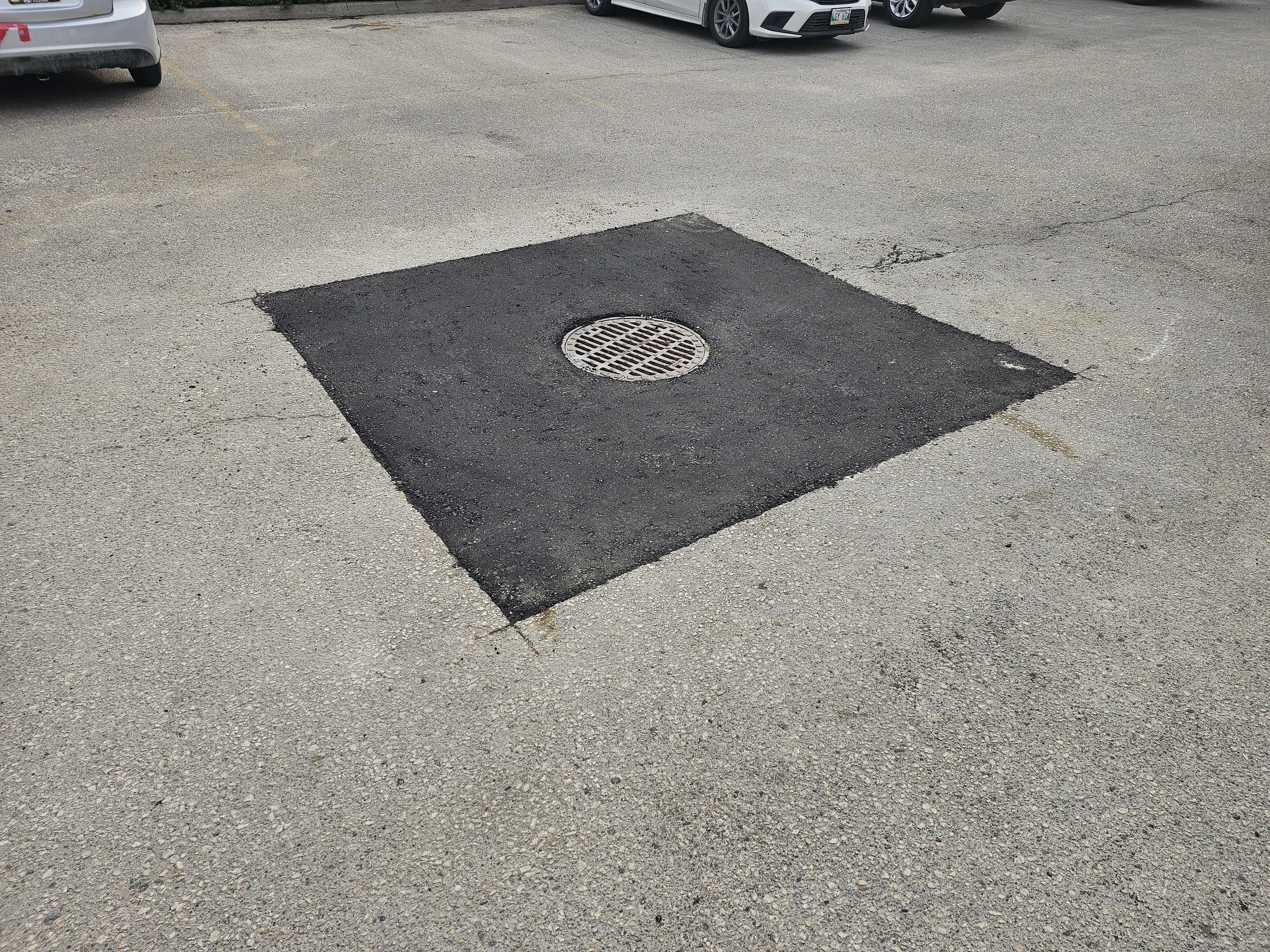 A parking lot with a square section of freshly paved asphalt surrounding a round metal drainage grate.