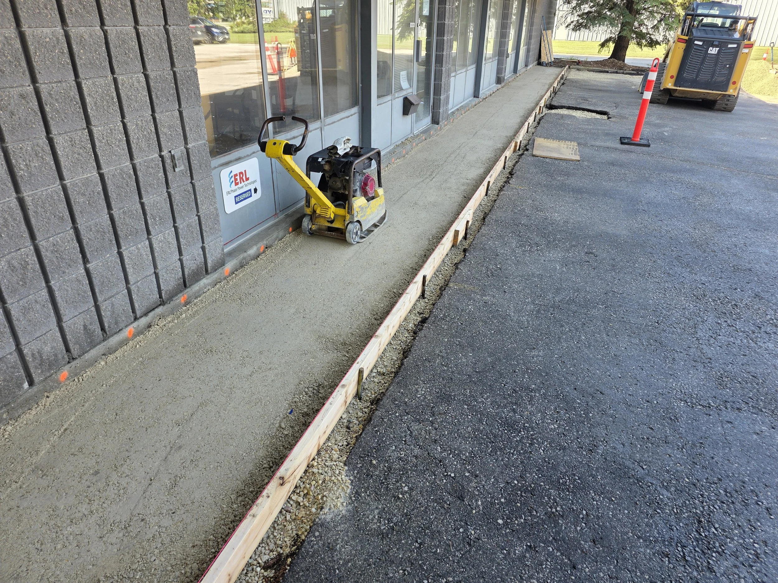 Construction site with freshly poured concrete sidewalk, a concrete screed machine, and red traffic cones, outside a building with glass windows.