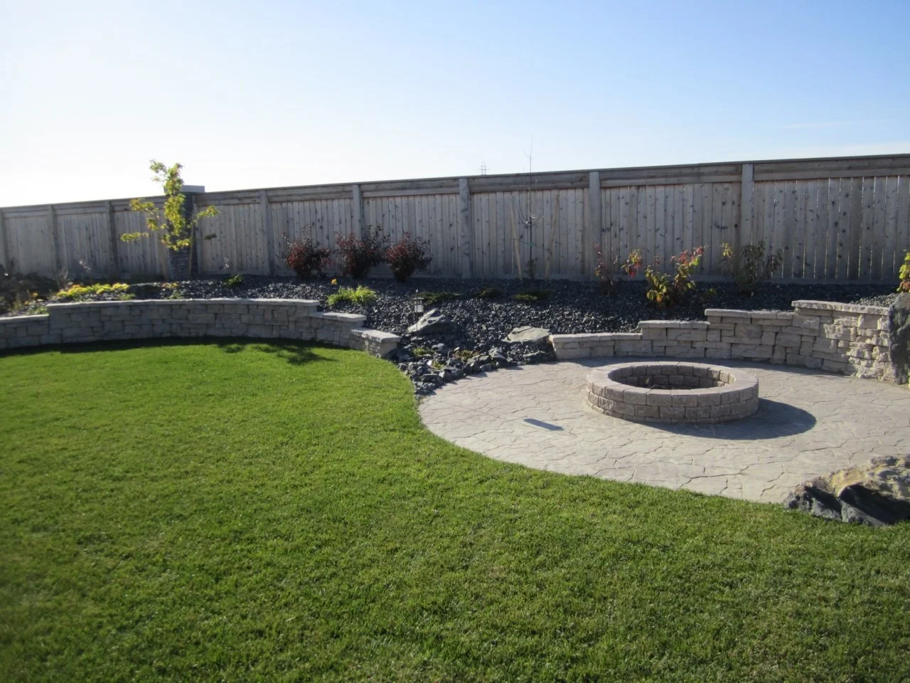 Backyard garden with a well-maintained green lawn, a circular brick fire pit, a paved patio area, and a raised garden bed with trees and shrubs, enclosed by a wooden fence.