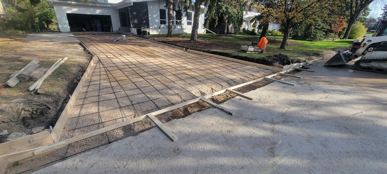 Construction workers are building a concrete driveway in front of a house. The area is in the process of being prepared with wooden forms and rebar, with a piece of construction equipment nearby.