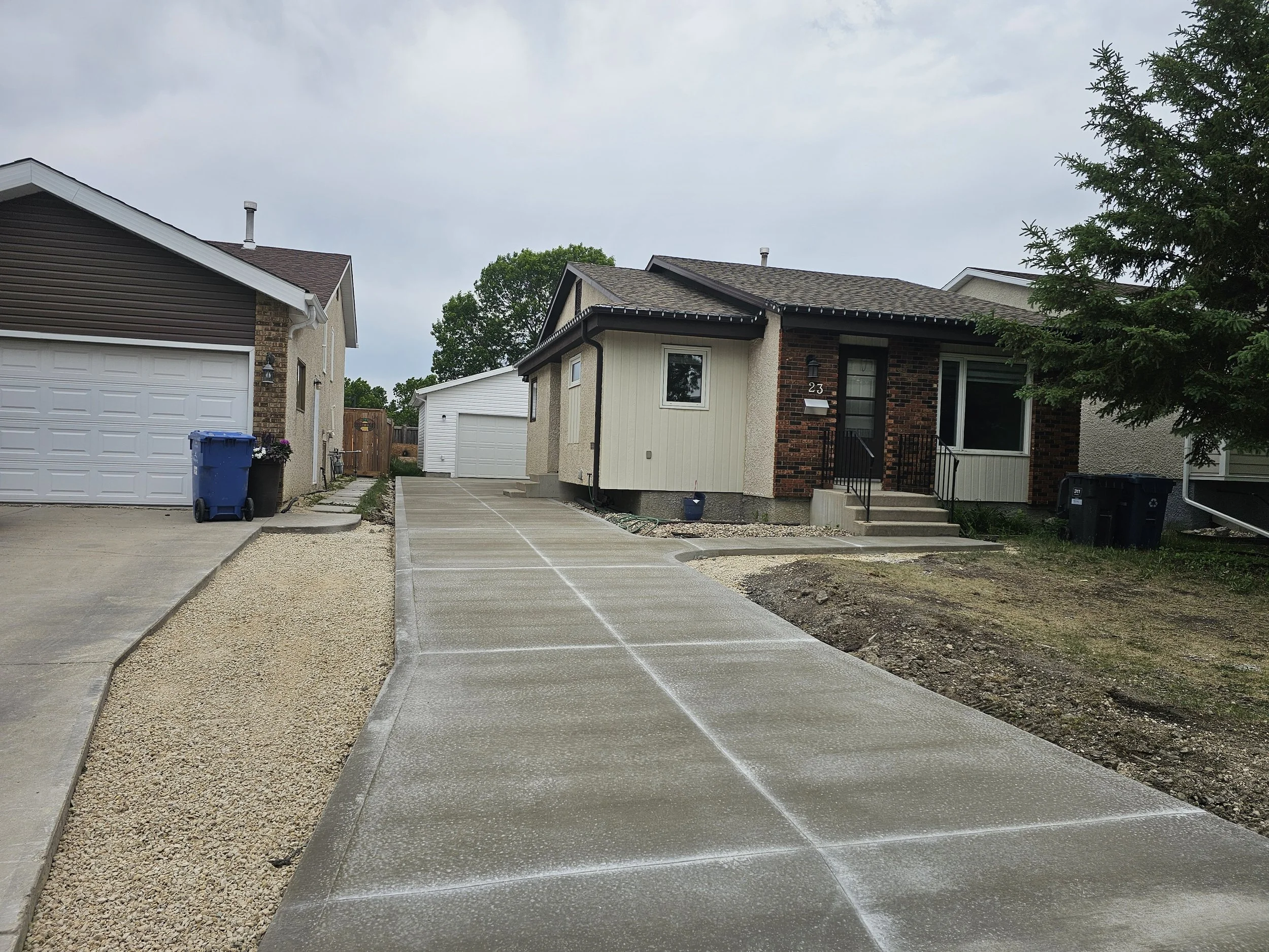 Driveway leading to the entrance of a single-story house with a brick and siding exterior, set in a residential neighborhood. The driveway has newly poured concrete and is flanked by gravel on one side. The house has steps leading up to the front door with black metal railings, and there are two black trash bins outside. The sky is overcast, and other houses are visible beside it.