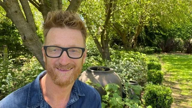 A man with glasses and a beard smiling outdoors near trees and bushes, with a large decorative pot behind him.