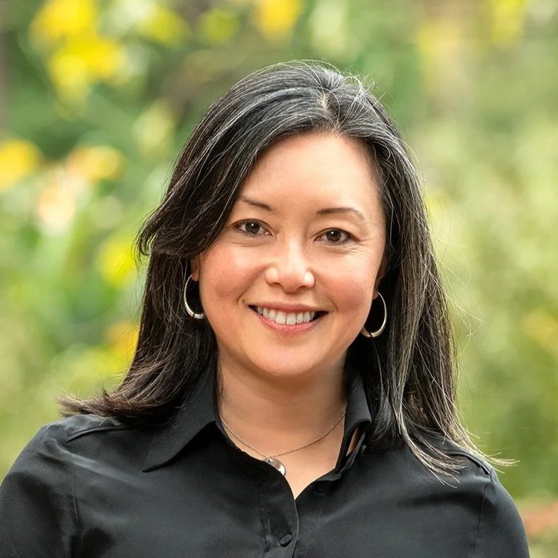 A woman with long dark hair and hoop earrings smiling outdoors with green and yellow blurred background.