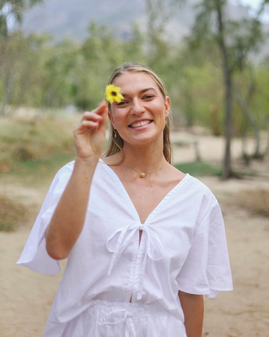 A woman smiling and holding a yellow flower outdoors with green trees in the background.