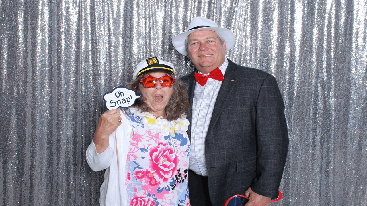 A woman and a man dressed in fun accessories posing in front of a silver, glittery backdrop. The woman is wearing a white jacket, a lei, red novelty glasses, and a captain's hat, holding a speech bubble prop that says "Oh Snap!" The man is wearing a gray checkered blazer, a white fedora, and a red bow tie, holding some party props.