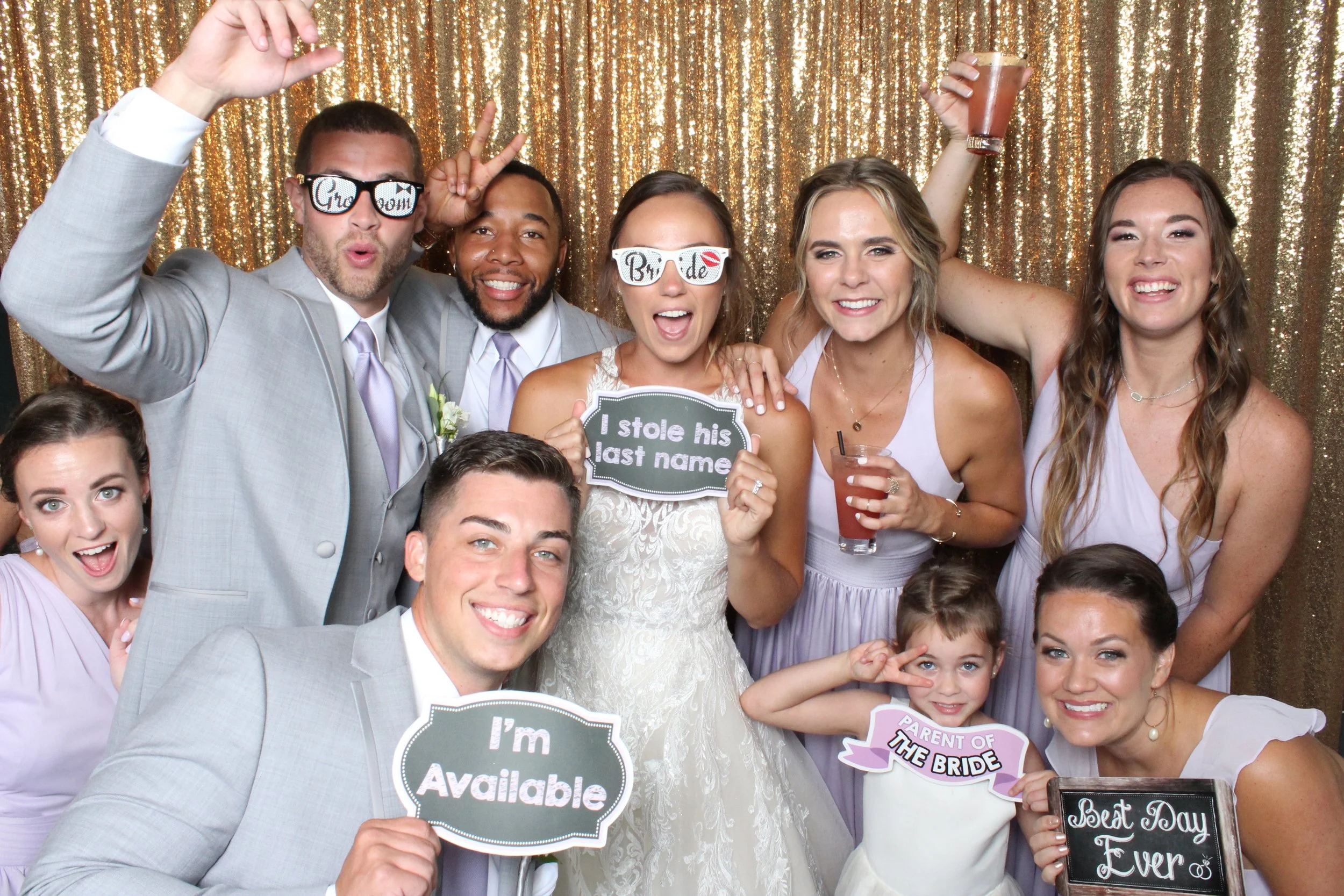 Group of people celebrating at a wedding with fun signs and party glasses in front of a gold glitter backdrop.