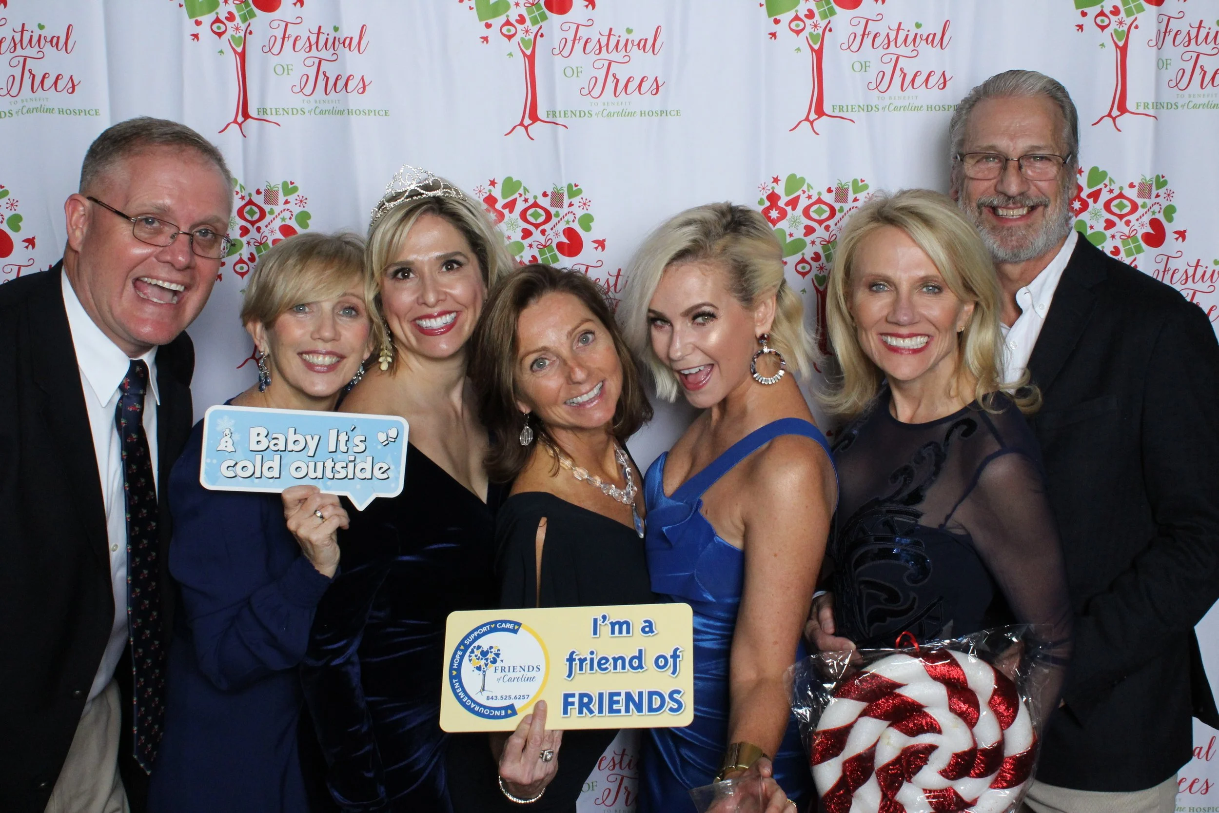 Group of seven smiling adults at a festive event, standing in front of a backdrop with Christmas tree and holiday decorations, holding signs that say "Baby It's cold outside" and "I'm a friend of FRIENDS."