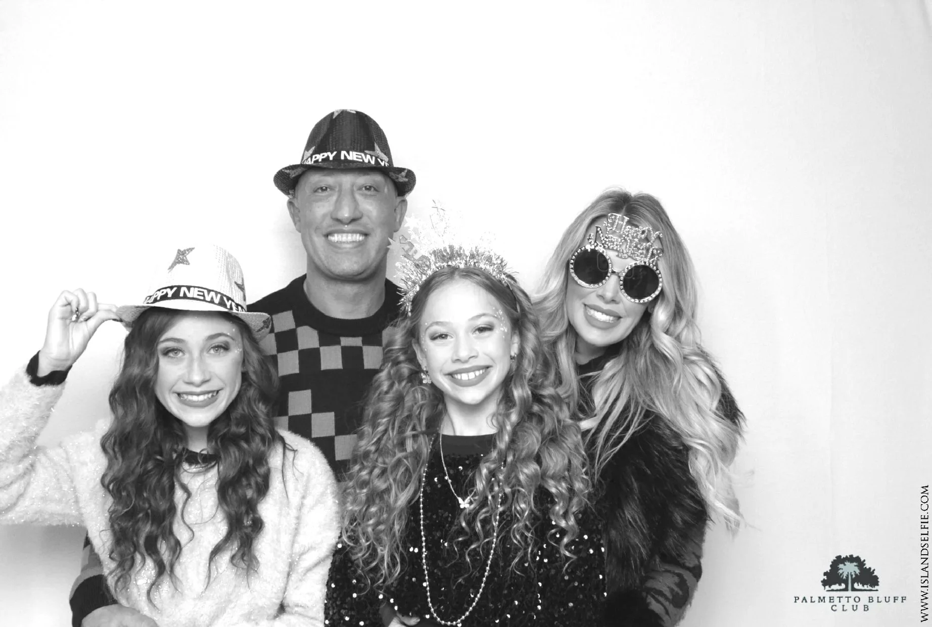 Group of four women and one man celebrating New Year's Eve, wearing hats and accessories with 'Happy New Year' themes, smiling for the camera, in a photo booth at the Palm Coast Civic Club.