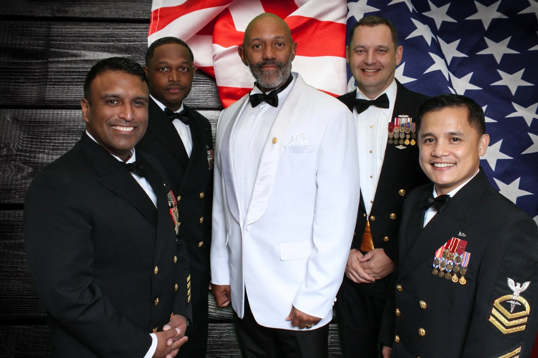 Group of five men dressed in military attire and a tuxedo, standing together in front of American flags and a wooden background, smiling at the camera.