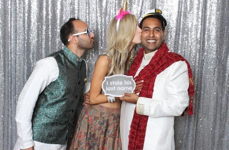 Three people in party costumes, with a woman kissing a man on the cheek, holding a sign that says 'I stole his last name', against a silver sequin backdrop.