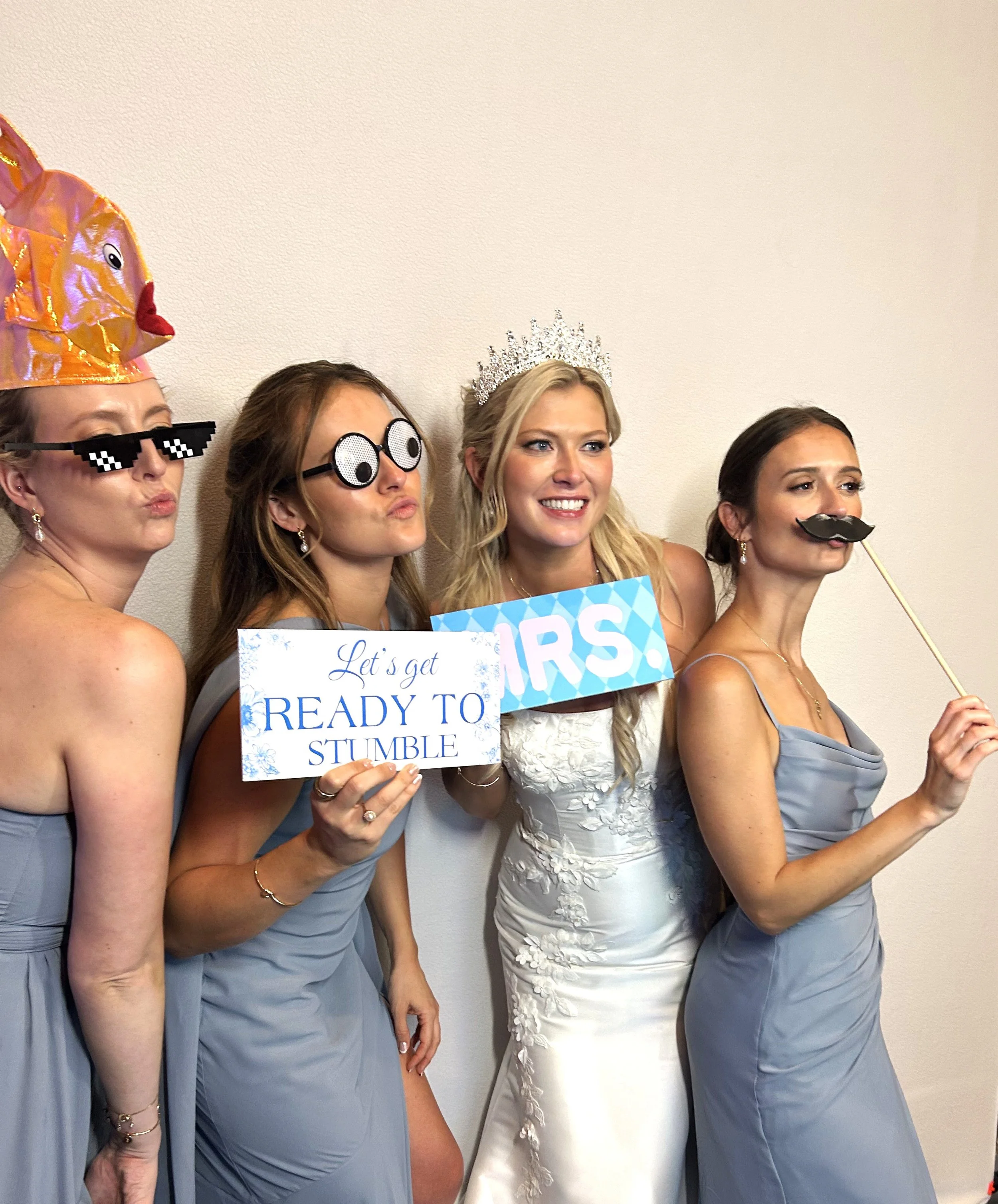 Four women dressing for a wedding, wearing blue dresses with one in a white wedding gown, holding glasses and signs, with one wearing a crown and another a party hat, posing against a plain wall.