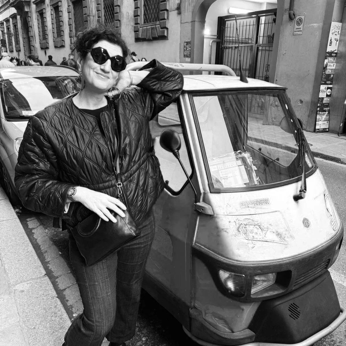 Rebecca Bedrossian, editor and co-founder of Curious Outpost. A woman with dark, curly hair wearing sunglasses and a quilted leather jacket leans against a small, three-wheeled vehicle parked on a city street.