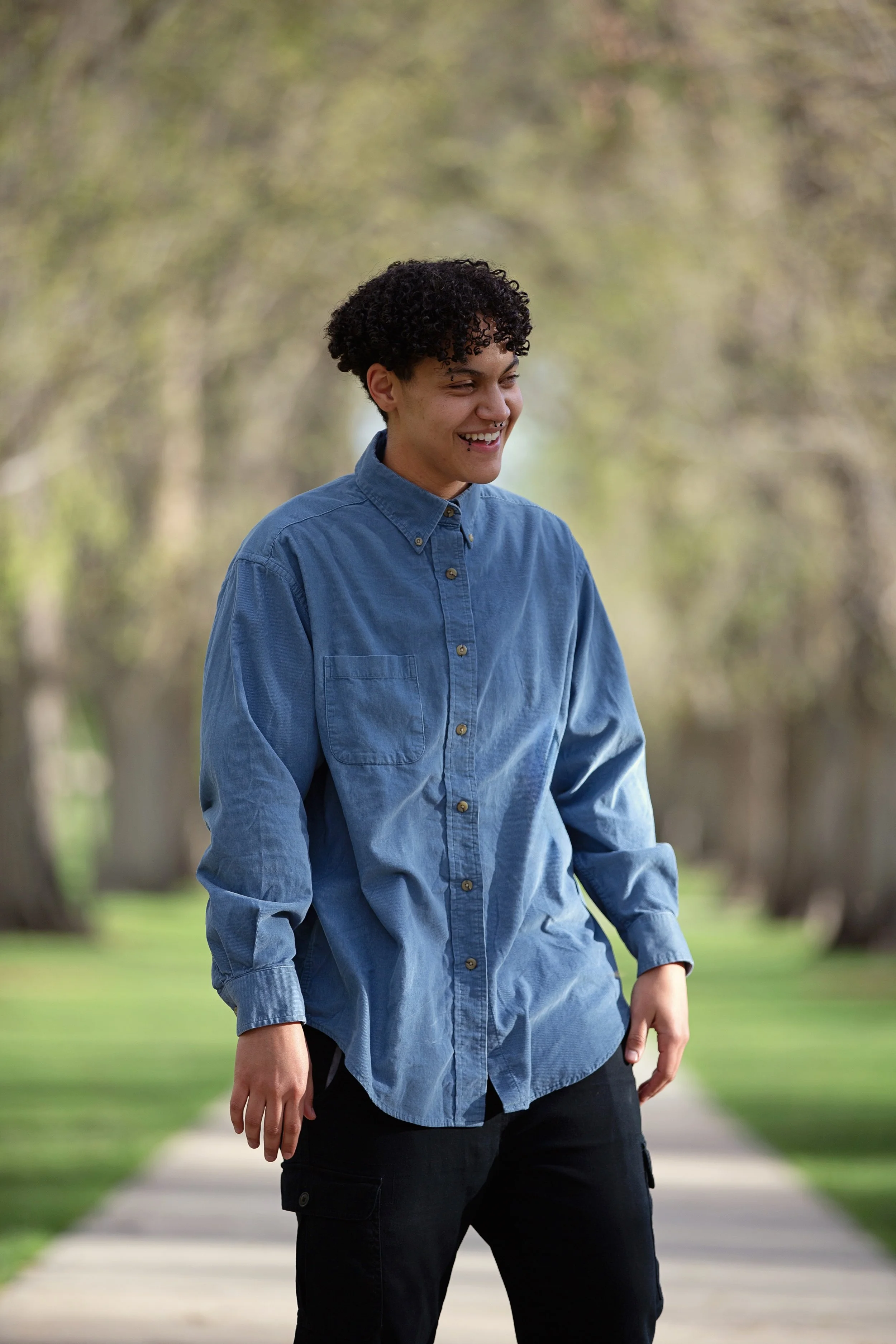 Photo of person laughing outdoors, framed above the knee, facing 45º away from camera. Person wears blue button down shirt and black pants.