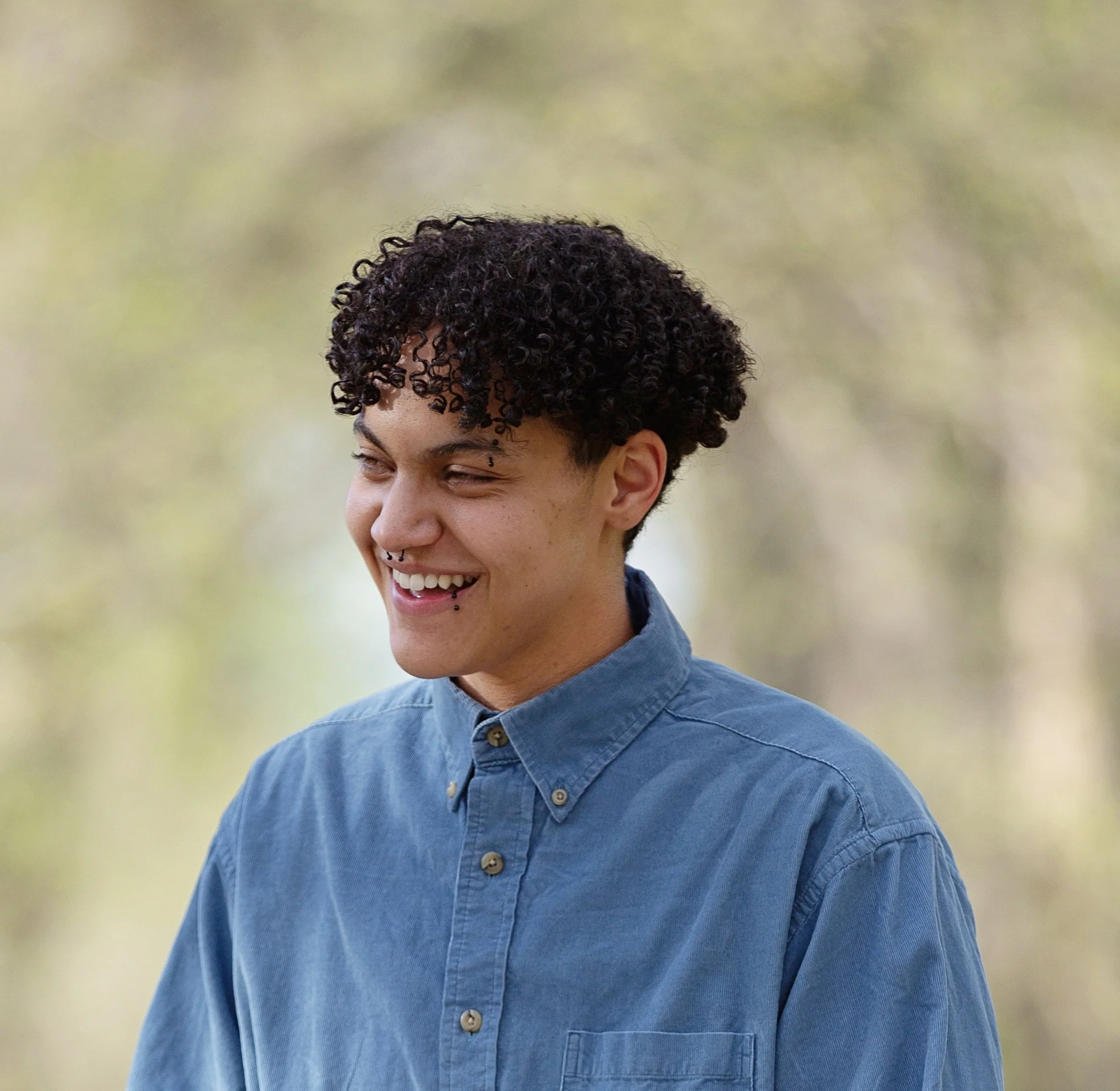 Close up headshot of Be Woods, above the chest. Blue button down shirt, person faces 45º away from camera laughing.