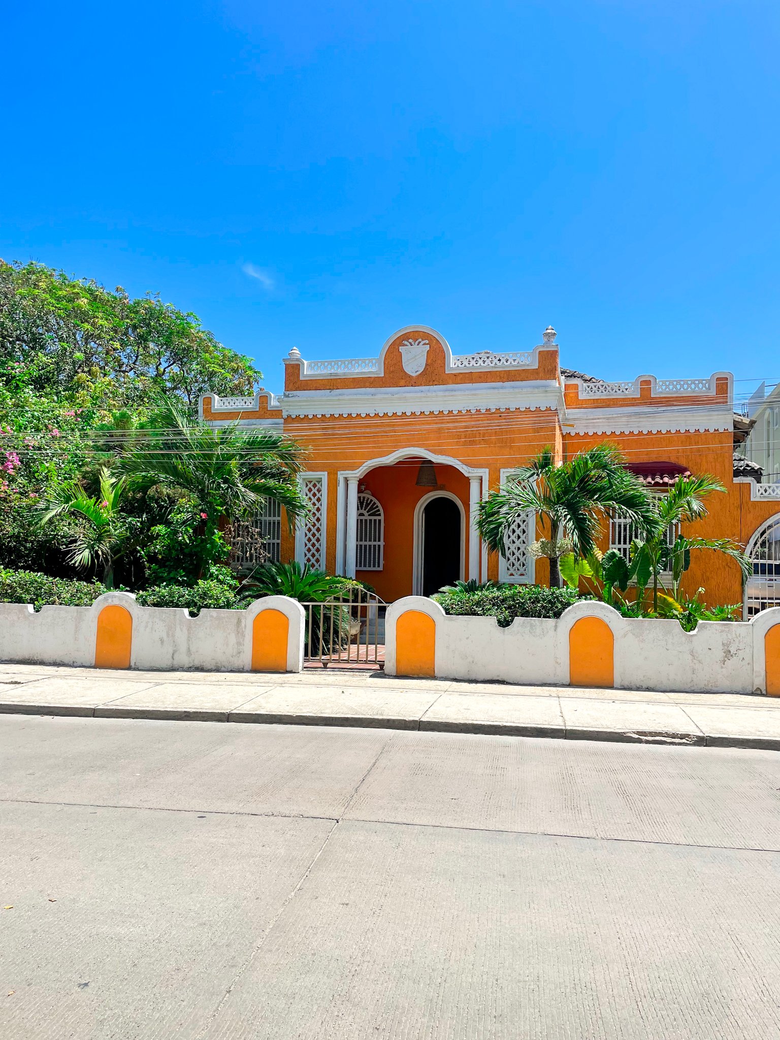 Orange house with white trim, decorated with a shield emblem with plants in front, separated from the sidewalk by a white fence with orange accents, under a bright blue sky.