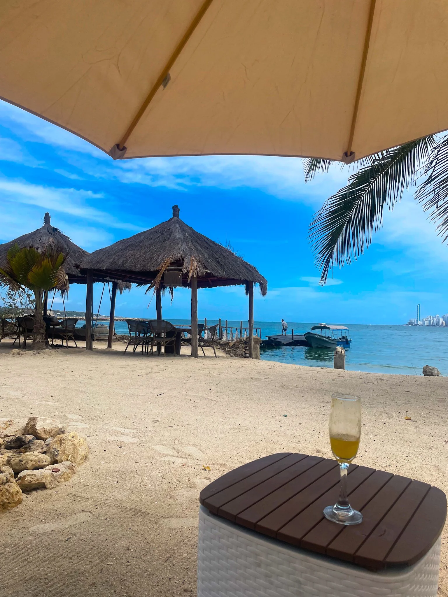 Beach scene with thatched umbrellas, wooden chairs, a palm tree, and a boat docked at the shore. In the foreground, there is a table with a wicker-patterned container and a glass of beer. City buildings can be seen in the distance on the right.