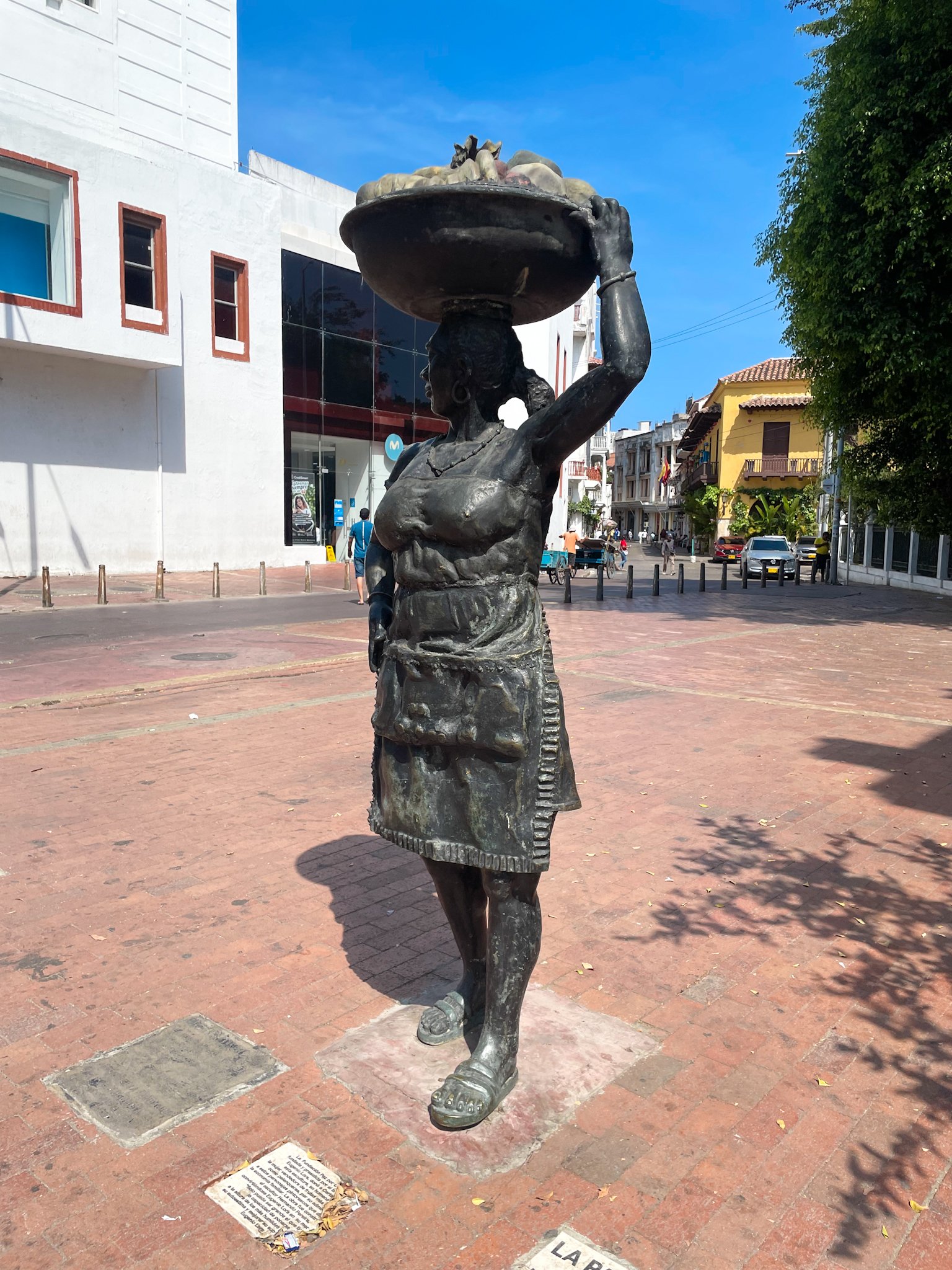 Bronze statue of a woman carrying a bowl on her head in a city street with buildings, trees, and cars in the background.