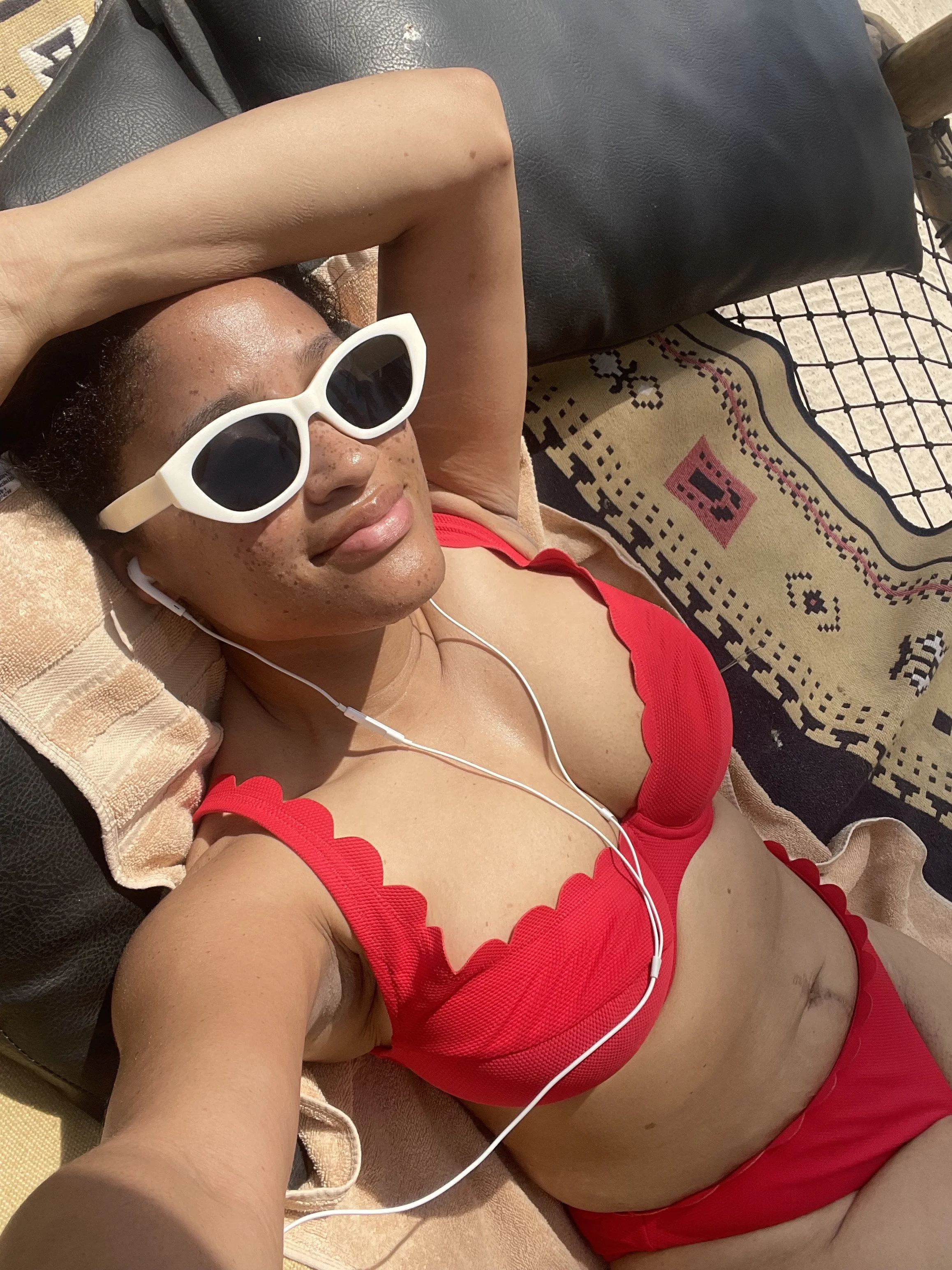 Person lying on a lounge chair outdoors, wearing white sunglasses, a red swimsuit with scalloped edges, and white earphones, with a towel underneath and a patterned rug nearby.