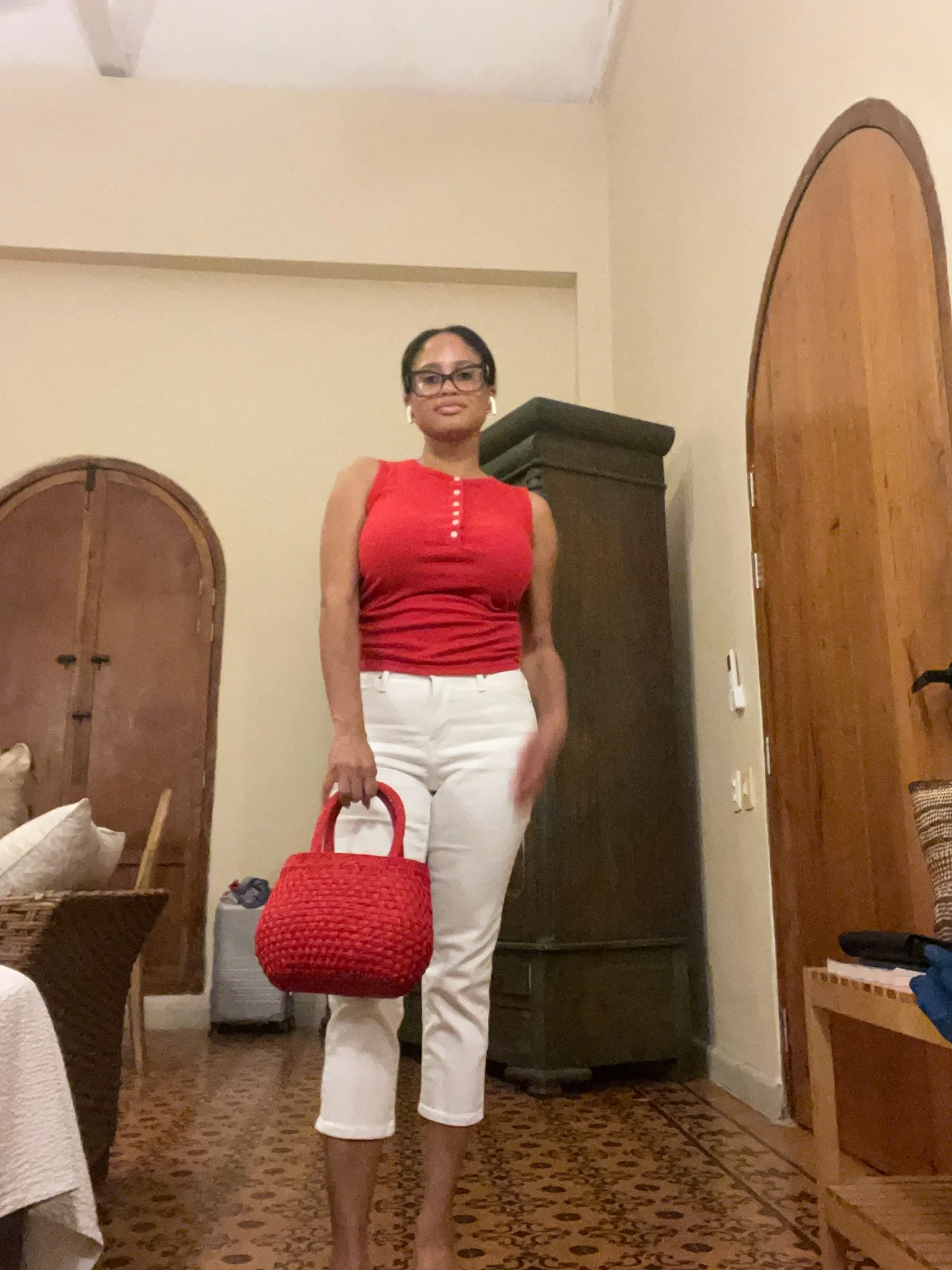 Woman in a red sleeveless top and white pants standing in a room, holding a red woven handbag. She has short dark hair, large glasses, and white earrings.
