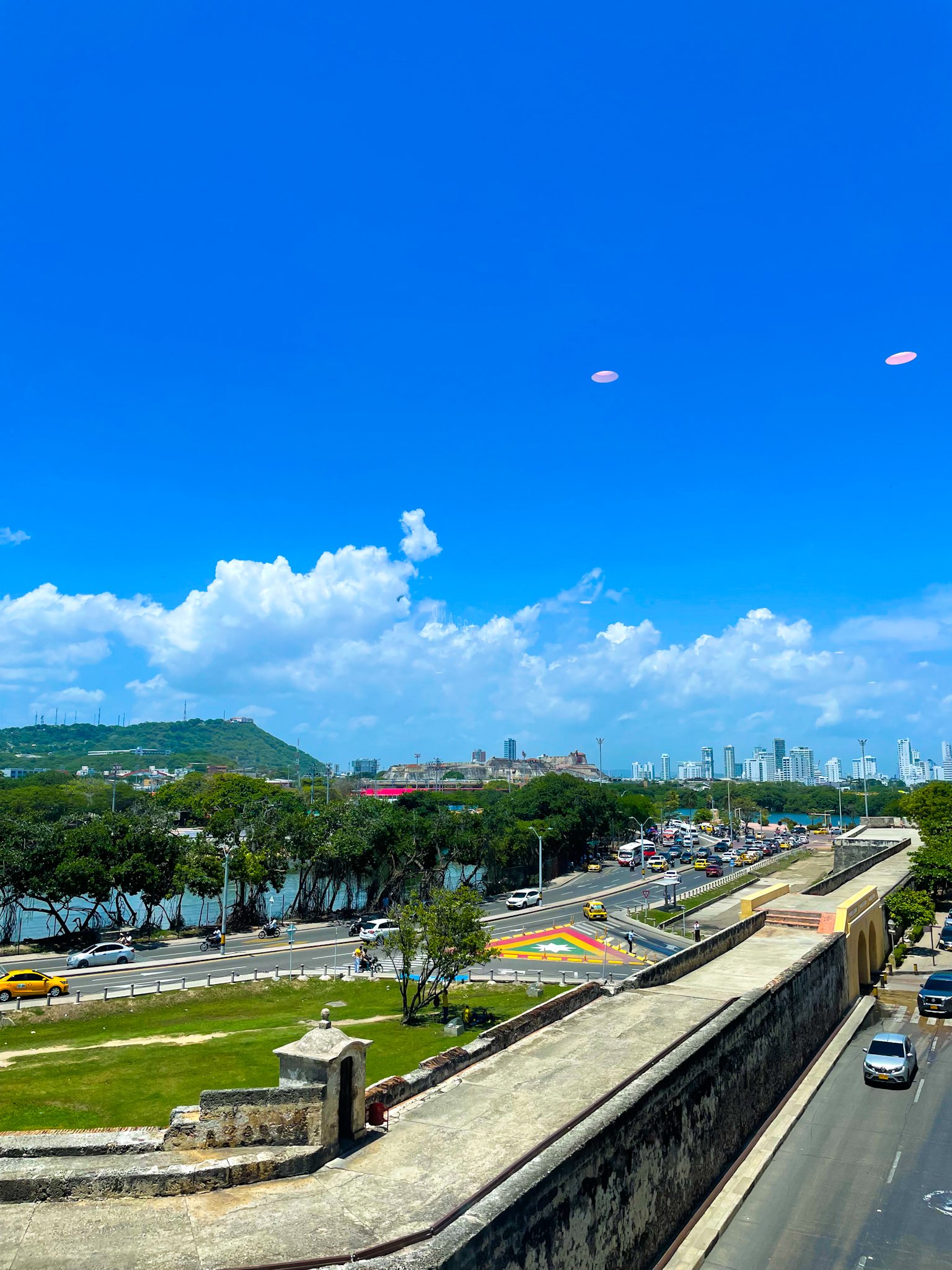 Cityscape with a highway and a park featuring a colorful geometric design, buildings in the distance, cloudy sky, and some trees.