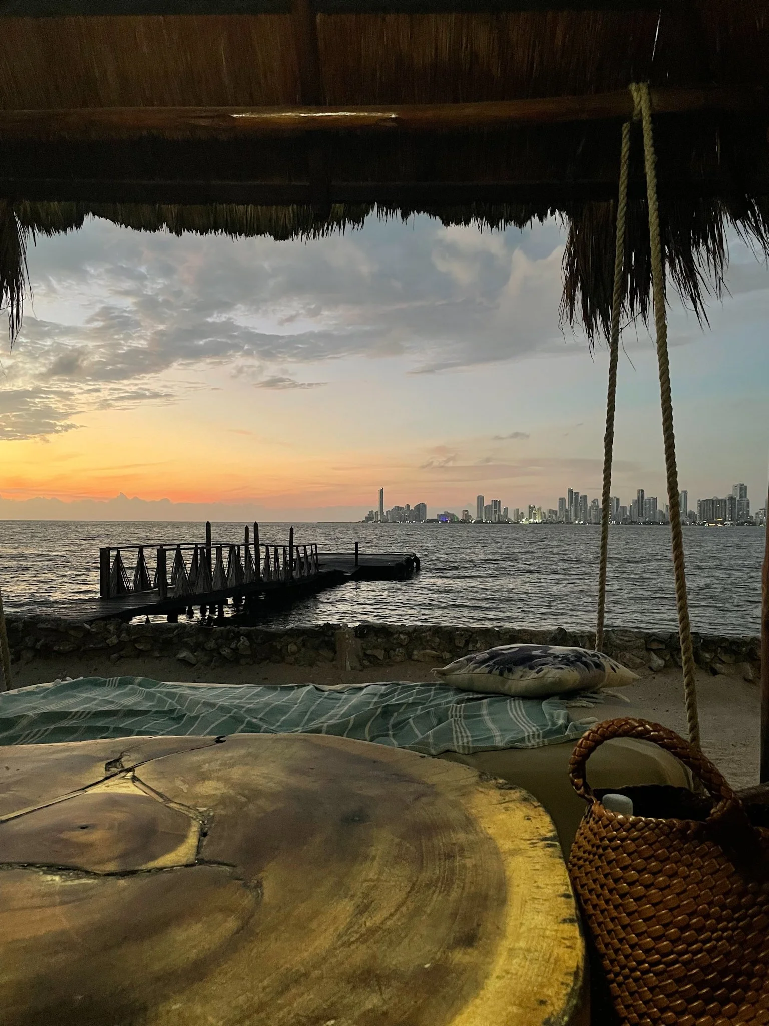 View from inside a thatched-roof hut overlooking the beach at sunset, with city skyline in the distance. There is a hammock with a pillow and a woven basket in the foreground.
