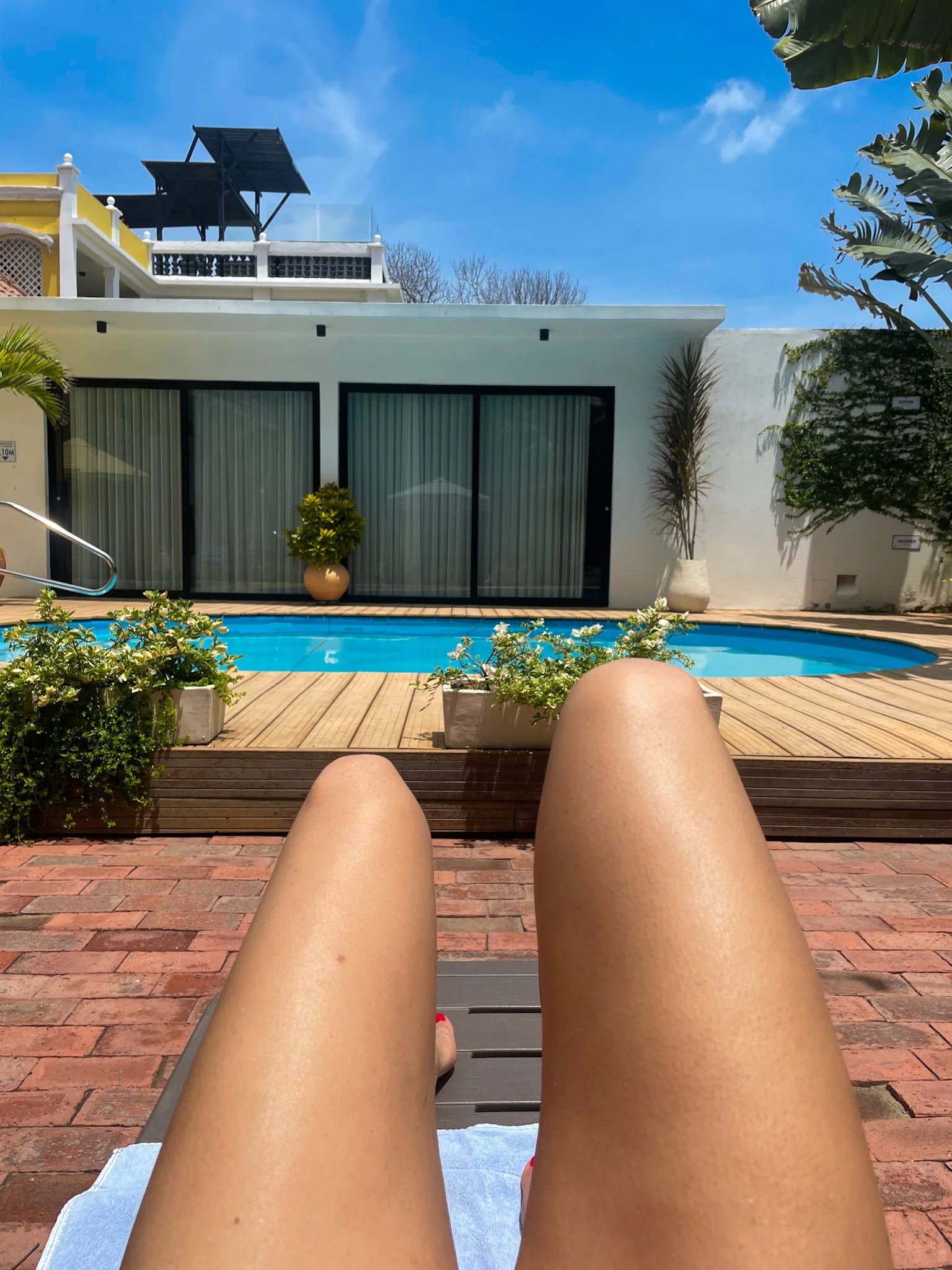 Person relaxing by a swimming pool on a lounge chair during a sunny day, with their legs extended in the foreground, in a backyard with potted plants and a modern white building with large glass doors.