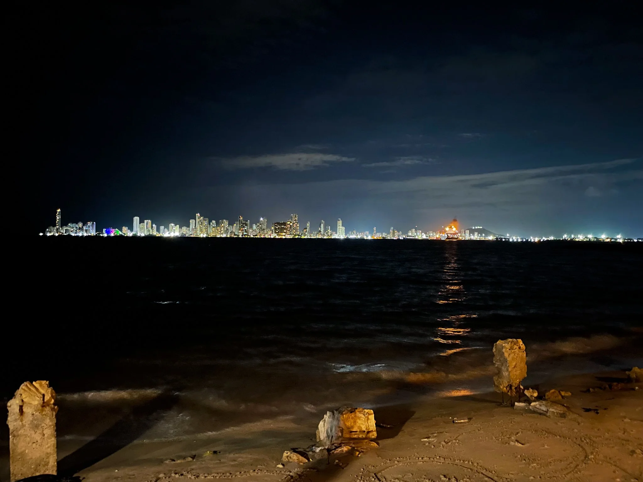 Night view of a city skyline with illuminated buildings across a body of water, seen from a beach with rocks and rocks on the shore.