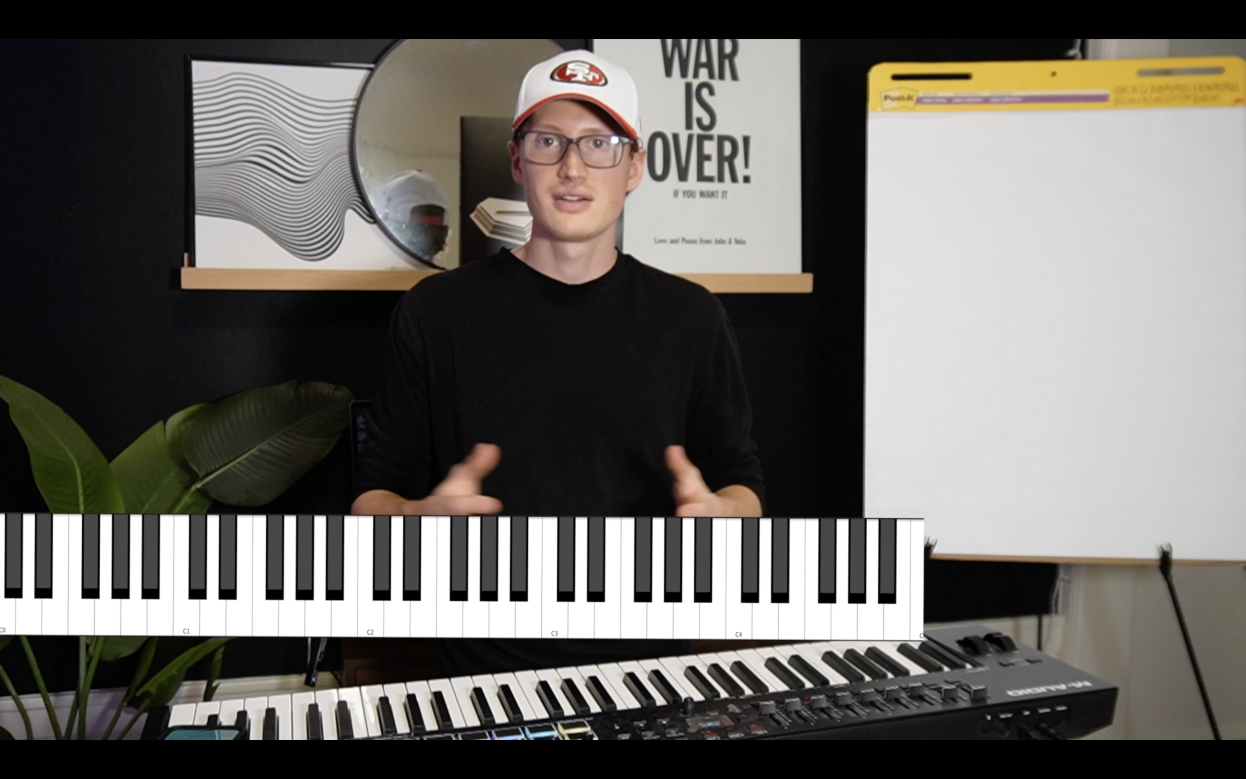 A young man with glasses and a red and white cap stands in front of a keyboard in a room with a black wall and decorative art. There is a whiteboard to his right and a poster that reads "WAR IS OVER!" behind him. A plant with broad green leaves is in the lower left corner.