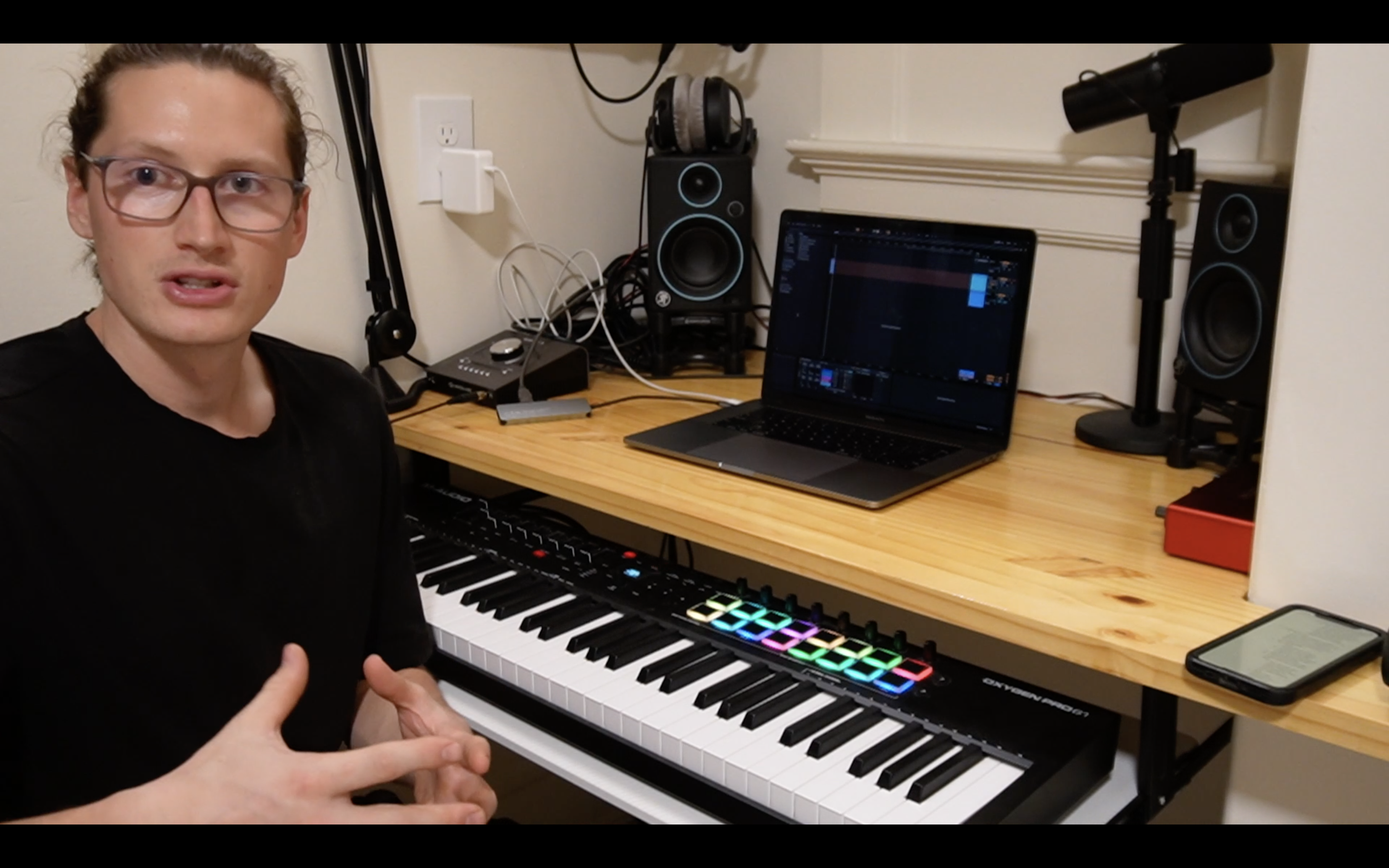 A young man with glasses and curly hair sitting at a keyboard setup, with a laptop displaying music production software, two studio monitor speakers, headphones, a microphone, and a smartphone on a wooden desk.