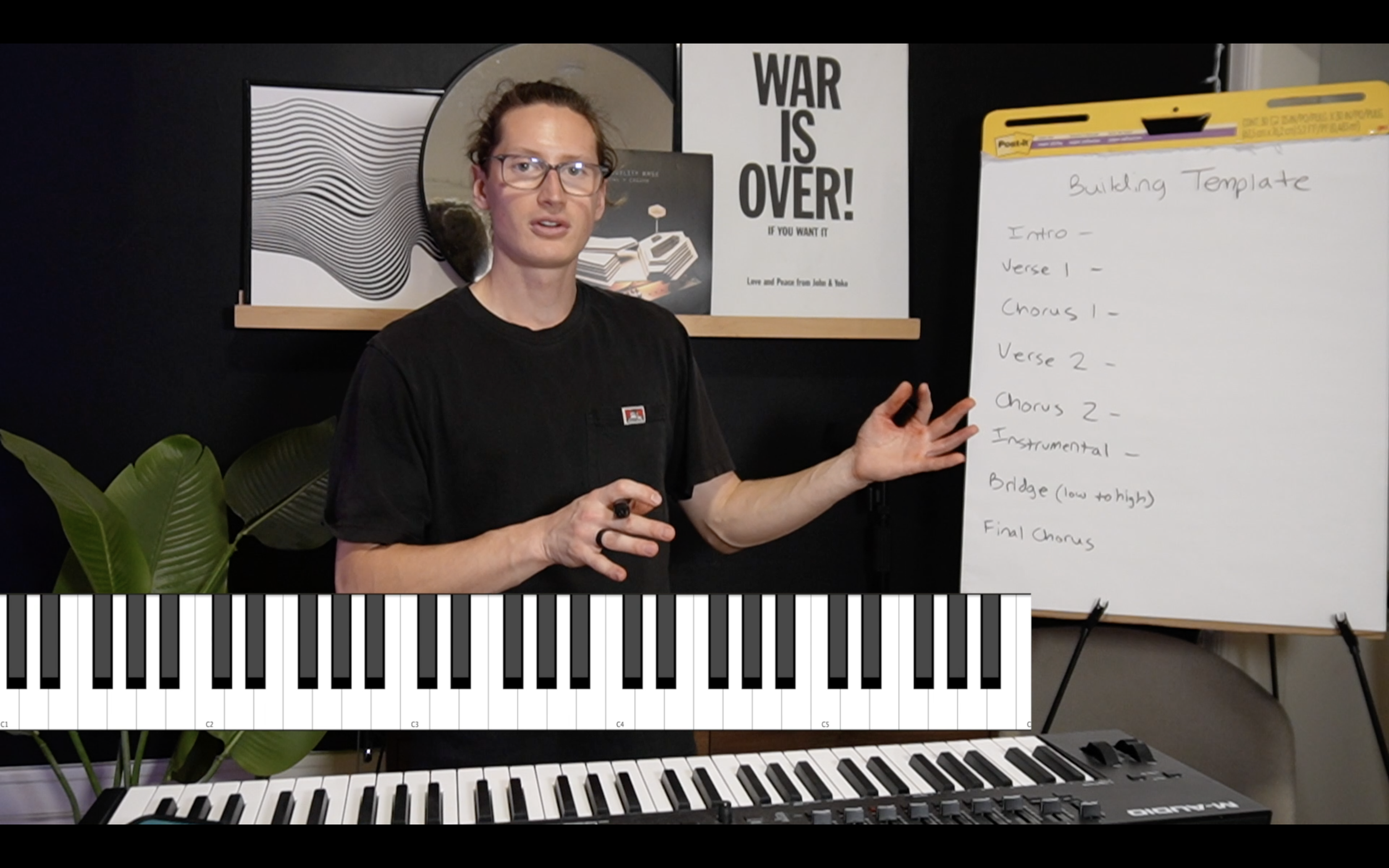 A person standing in front of a whiteboard with a music building template, next to a keyboard and a plant, with posters behind them including one that says 'WAR IS OVER!'