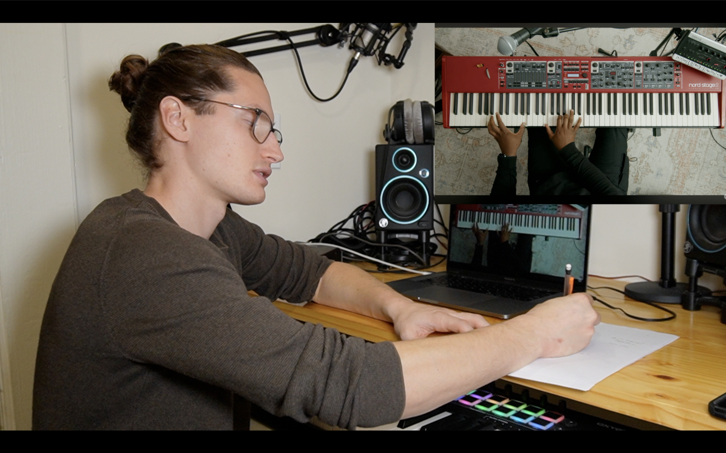 A person writing on a notepad at a music production workspace with a laptop, studio speakers, headphones, and a MIDI controller keyboard. On a monitor, a mirrored image shows the person playing the keyboard.
