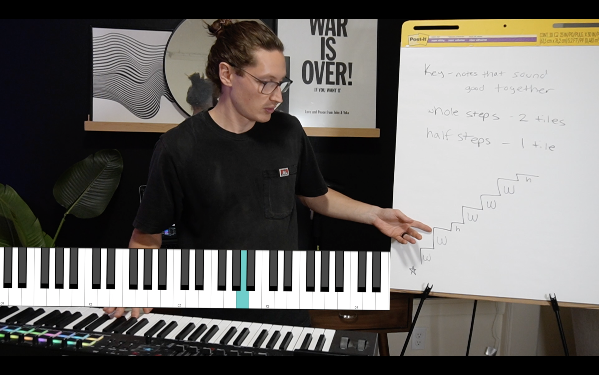 A man with glasses and long hair tied back, wearing a black T-shirt, is standing and pointing at a whiteboard with musical notes and steps written on it. There is a digital piano and keyboard in front of him, and a poster behind him with the text "WAR IS OVER!" and a black-and-white abstract art piece on the wall.