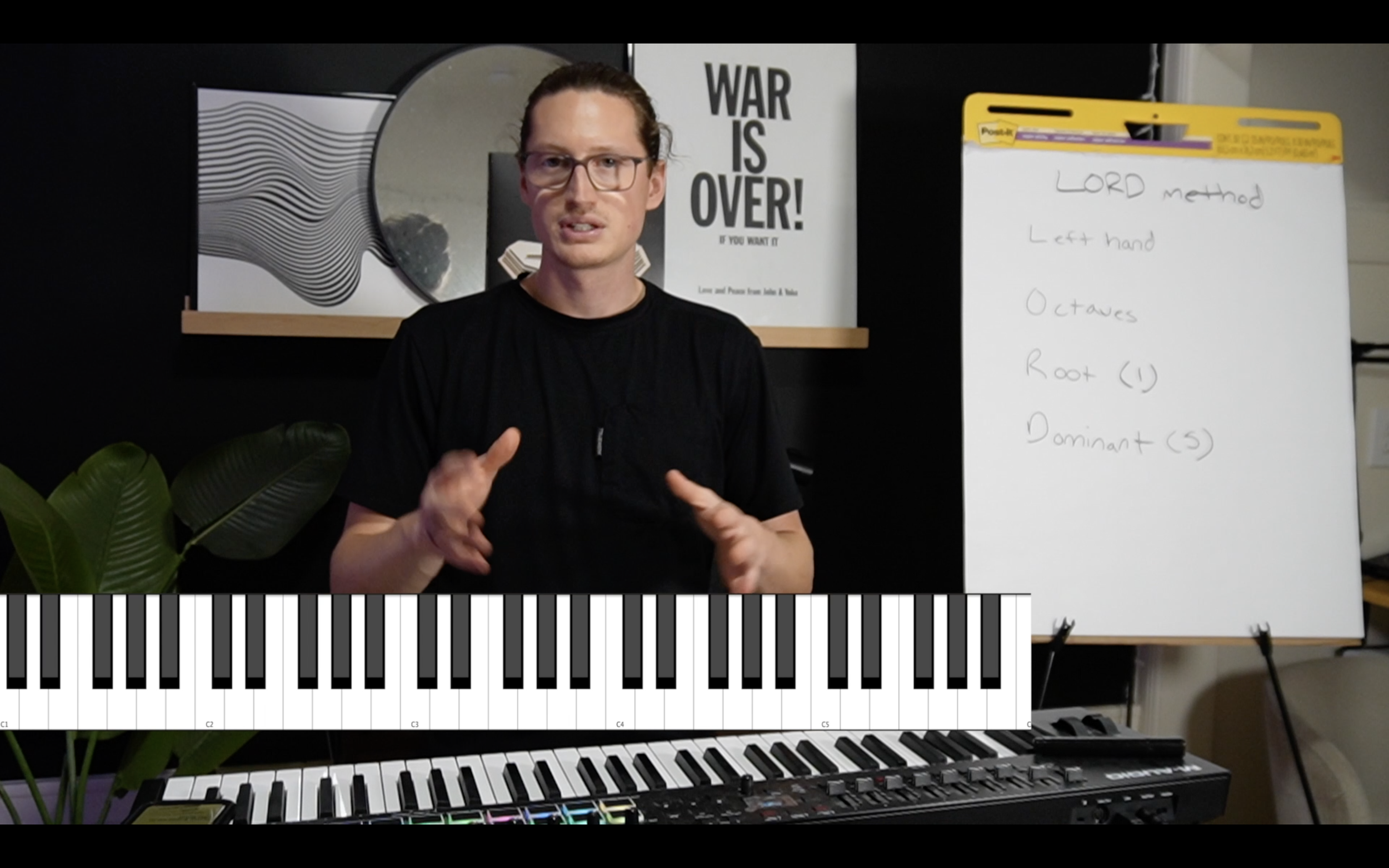 A person with glasses and long hair tied back, standing in a room with a keyboard and a whiteboard, gesturing with their hands. The room features a round mirror, a plant, and a poster that reads 'WAR IS OVER!'
