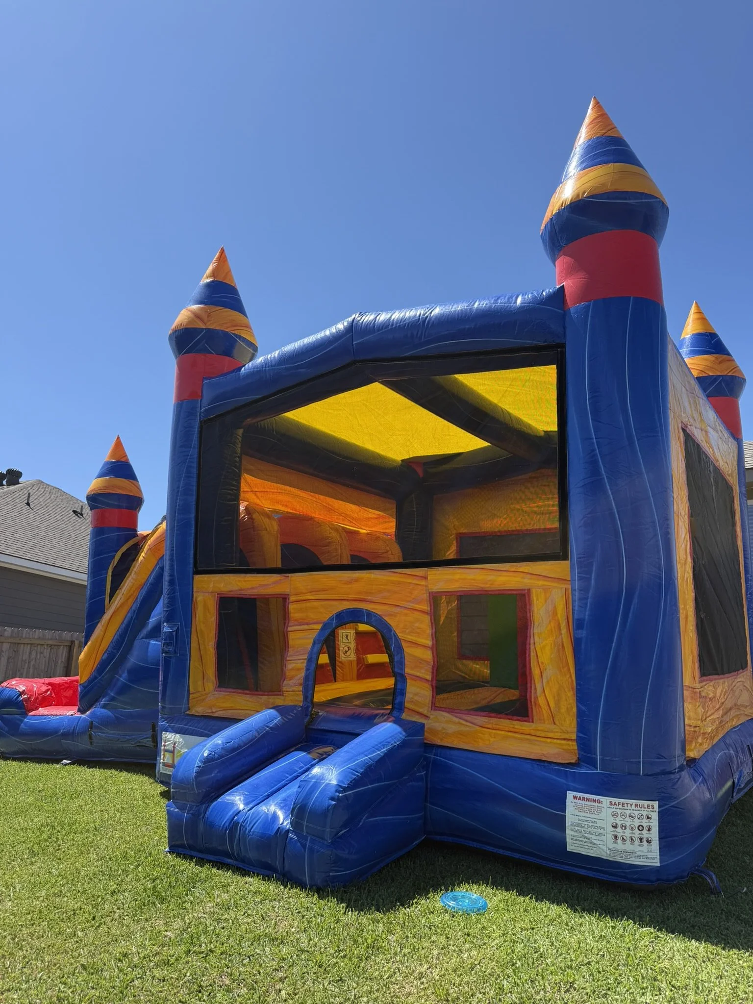 Colorful inflatable castle with turrets and a slide, set up on a grassy yard under clear blue sky.