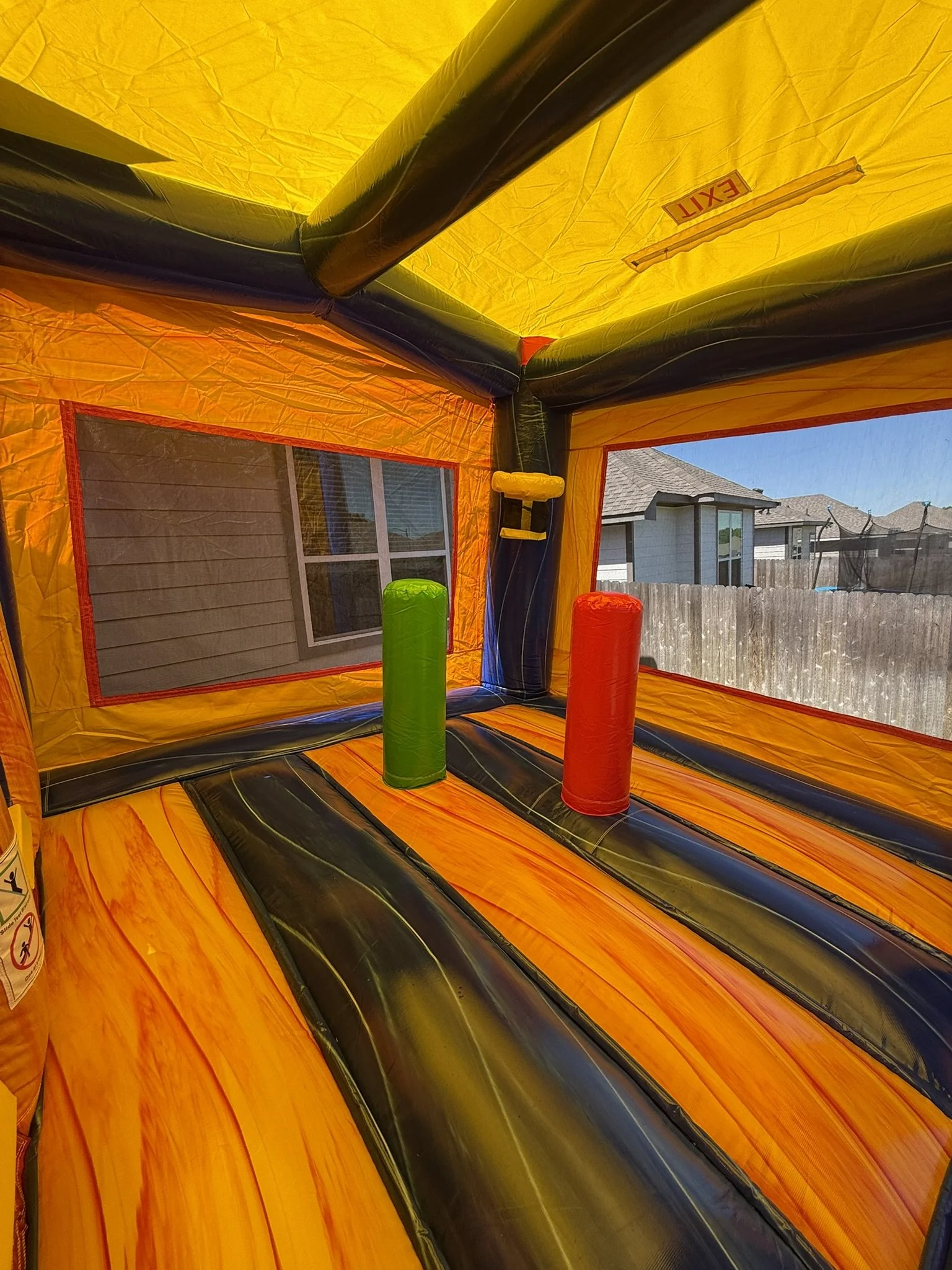 Inside view of a colorful inflatable bounce house with orange, yellow, black, green, and red colors, overlooking a suburban backyard with houses, a wooden fence, and a trampoline.
