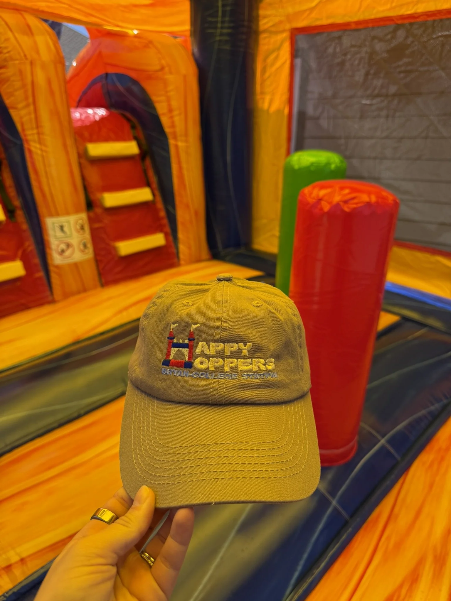 A tan baseball cap with the words 'Happy Hoop' embroidered on it, held in front of an inflatable bounce house with orange, yellow, red, green, and blue colors.