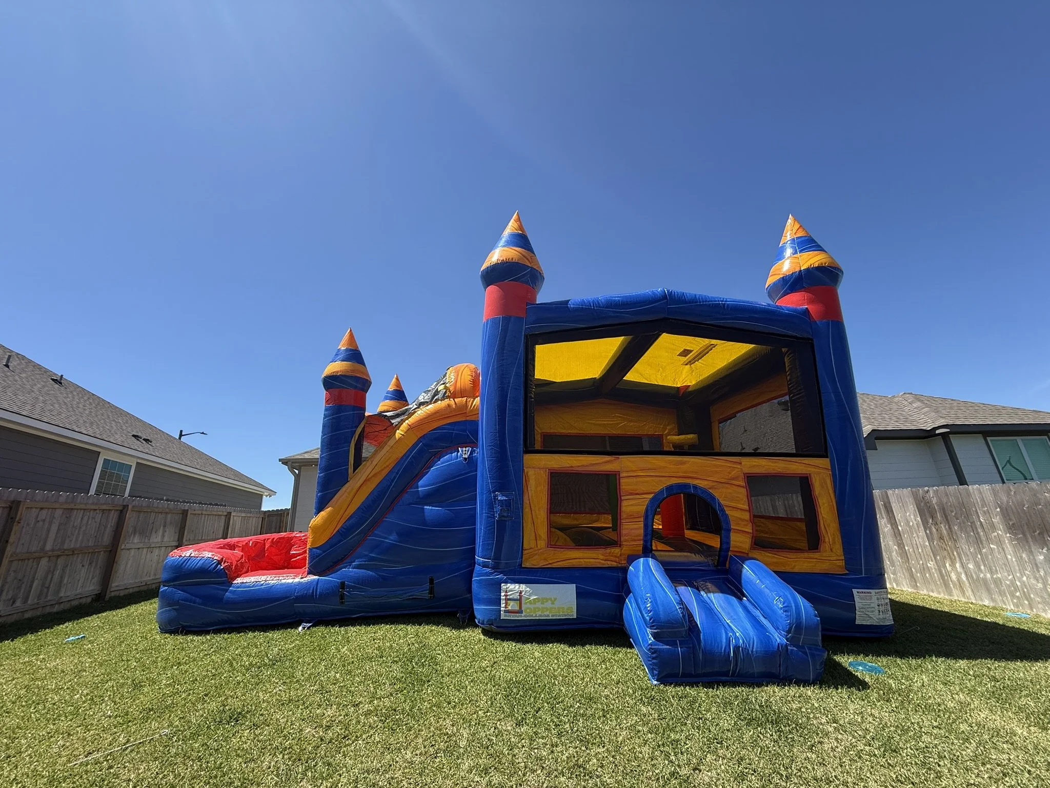 Colorful inflatable bounce house with a slide, designed to resemble a castle, set up in a backyard with green grass and wooden fences. The sky is clear and blue.