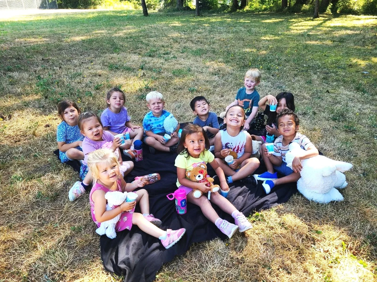 Children sitting on a blanket having a picnic outdoors in a grassy area with trees in the background, holding snacks and toys.