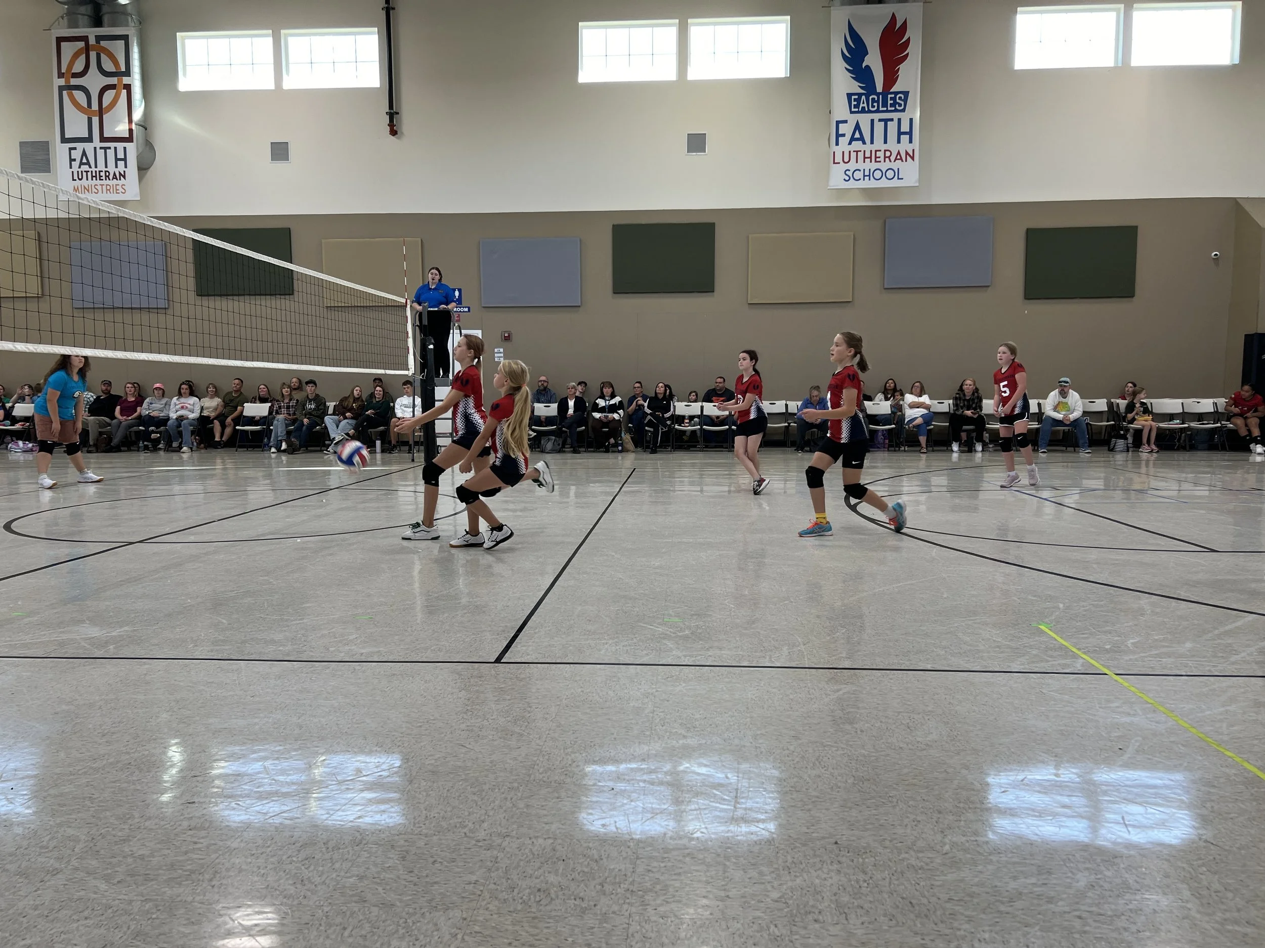 Indoor volleyball game with five girls in red jerseys on the court, one girl in a blue jersey serving the ball, and spectators seated along the wall. Team is playing at Faith Lutheran School, as indicated by banners hanging from the wall.